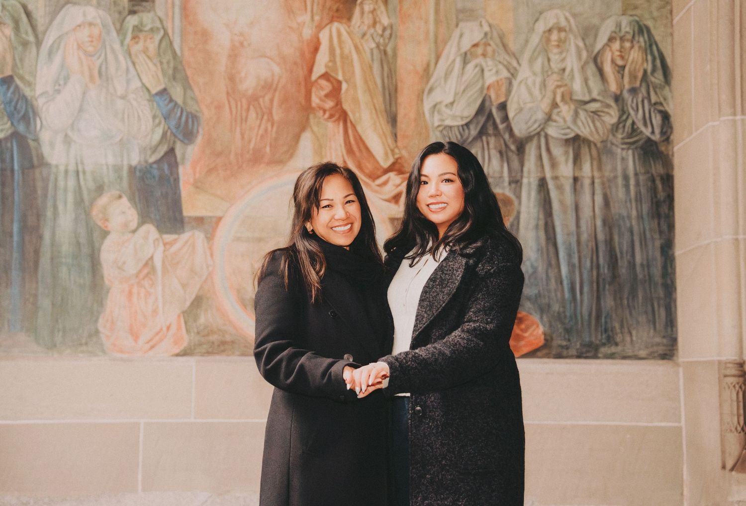 Mom and daughter posing together smiling in front of a large ornate religious wall painting.