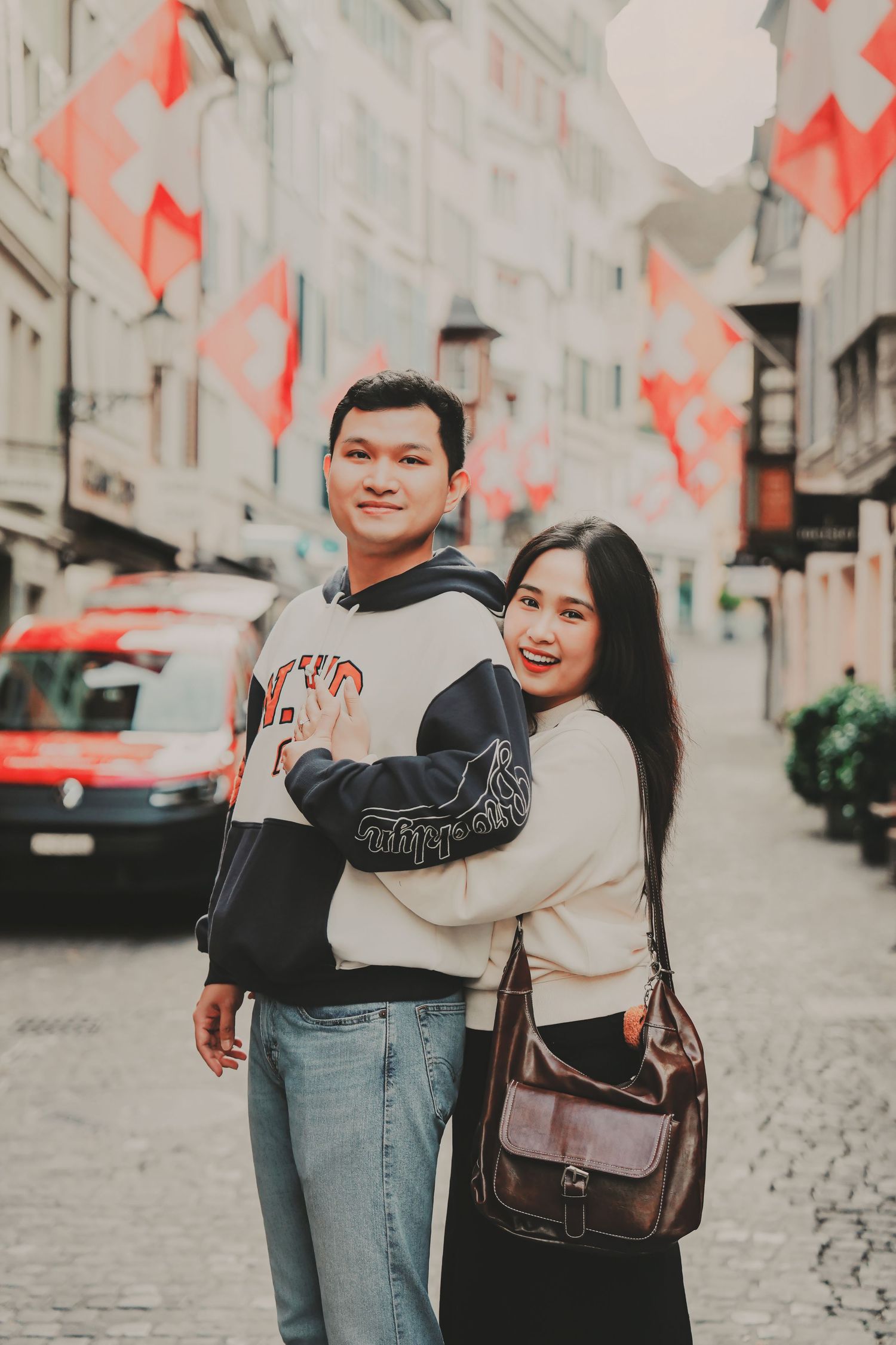 Happy Asian couple embracing on cobblestone street with Swiss flags in background in Zurich.