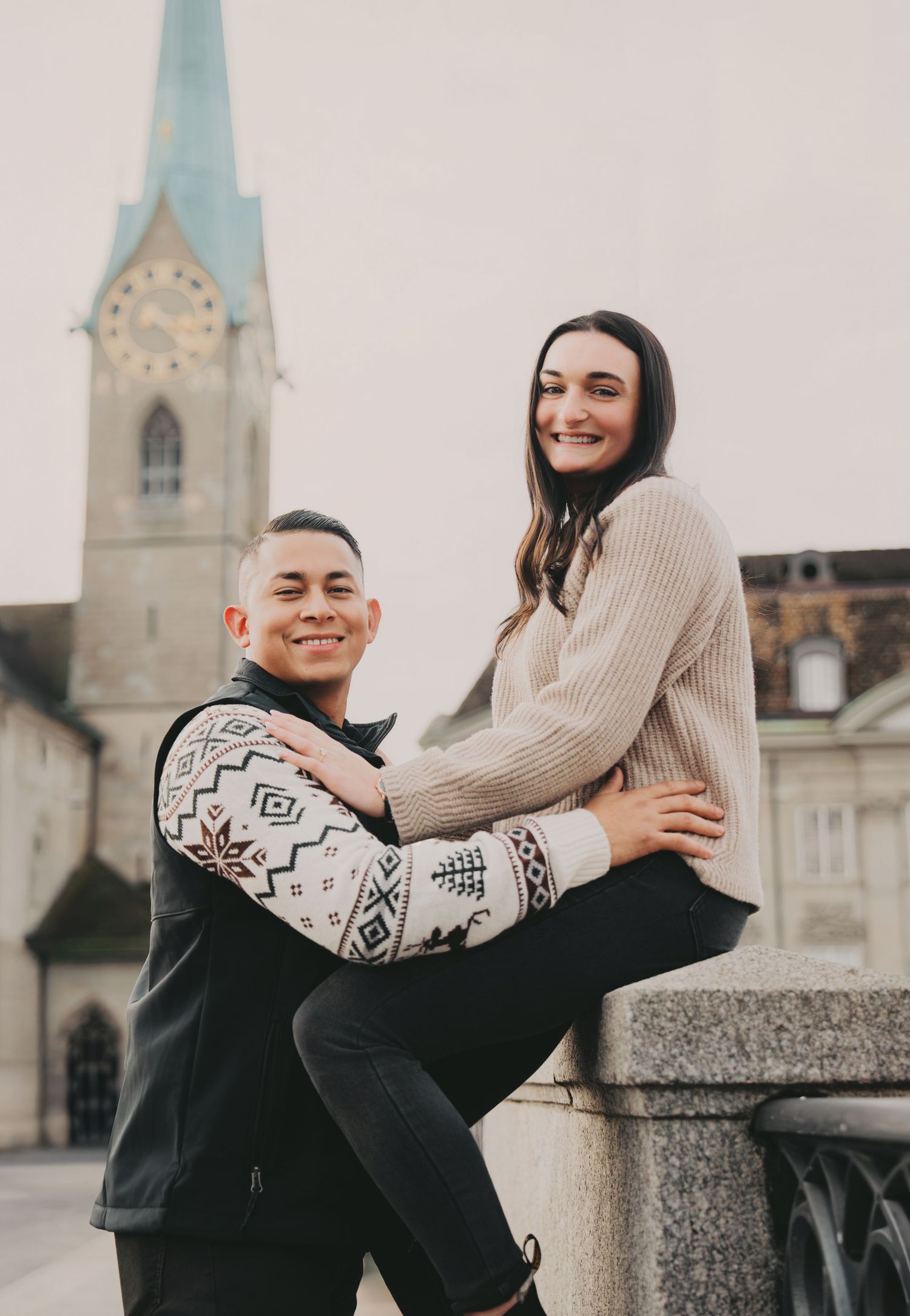 Smiling couple posing on a stone ledge with Fraumunster Church clock tower in Zurich behind.