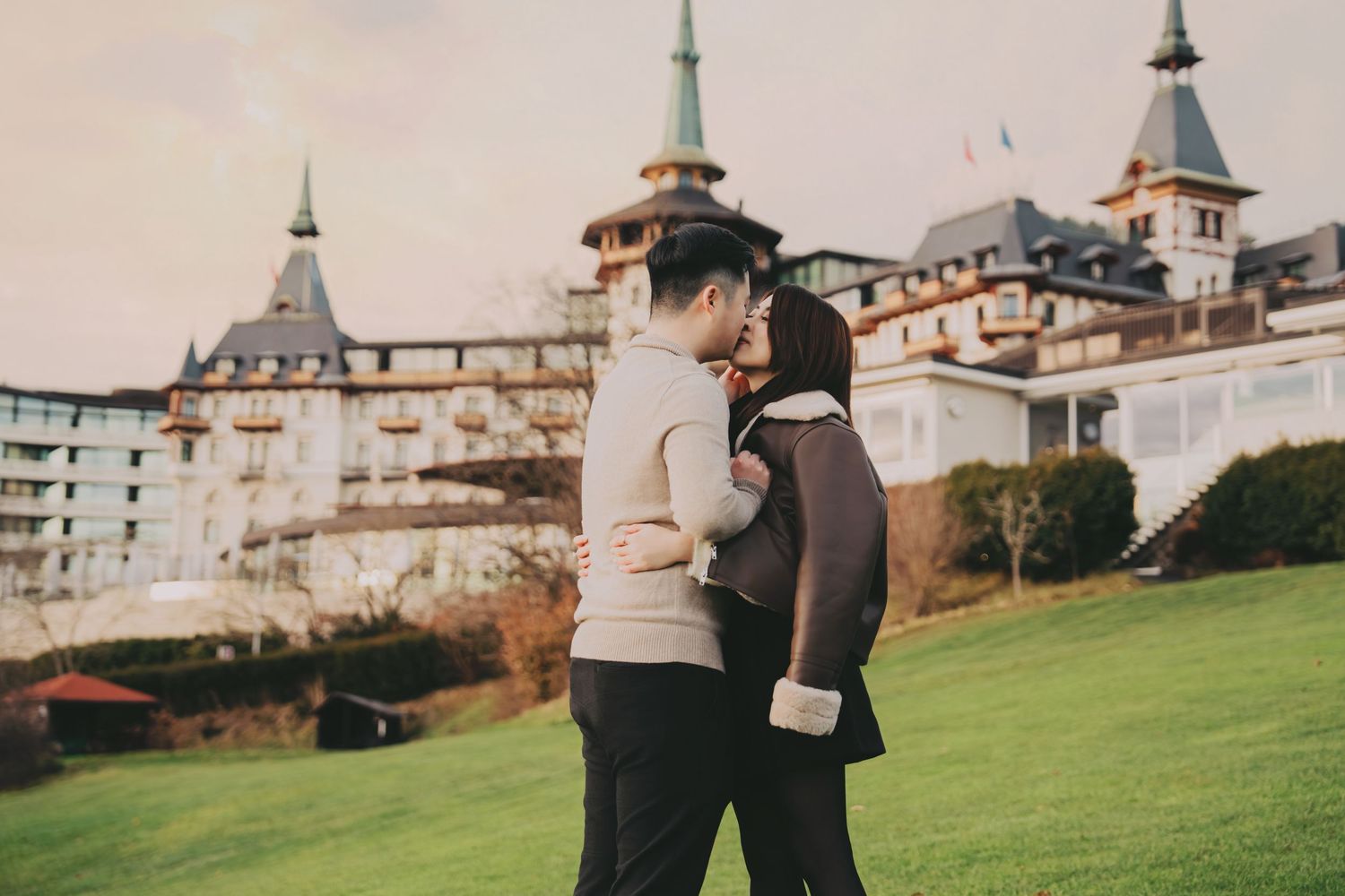 Asian couple sharing romantic kiss on grass with grand European château behind them.
