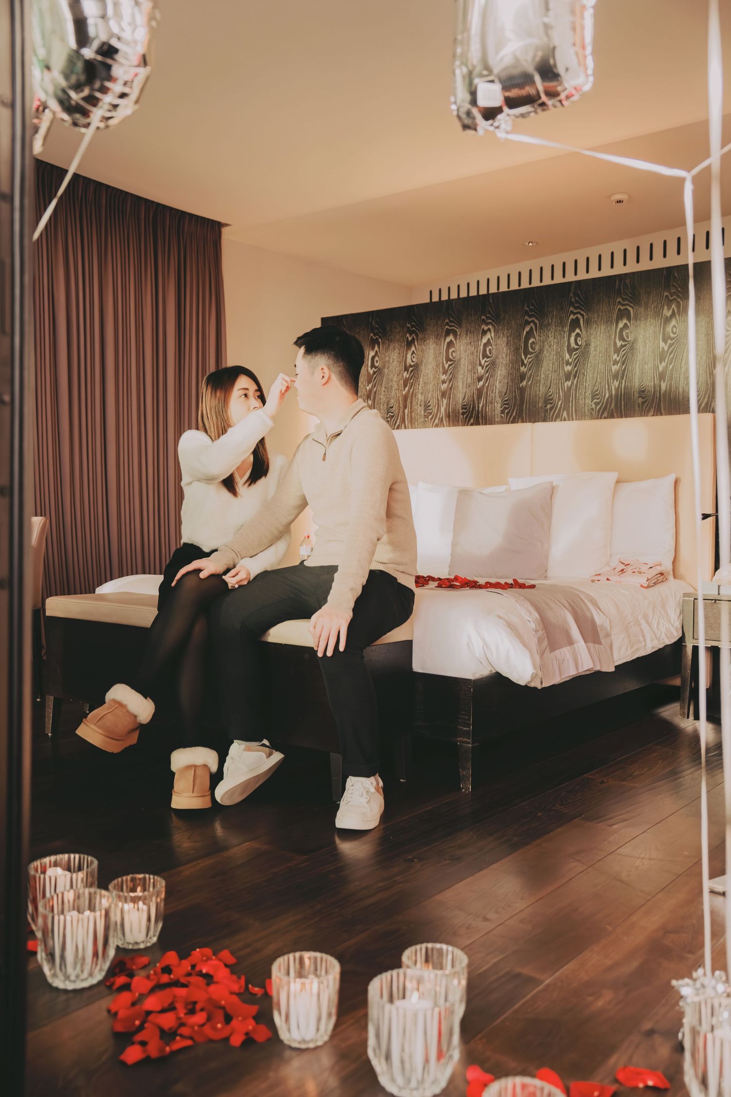 Romantic hotel room setup with rose petals and candles as Asian couple sits together on bed.