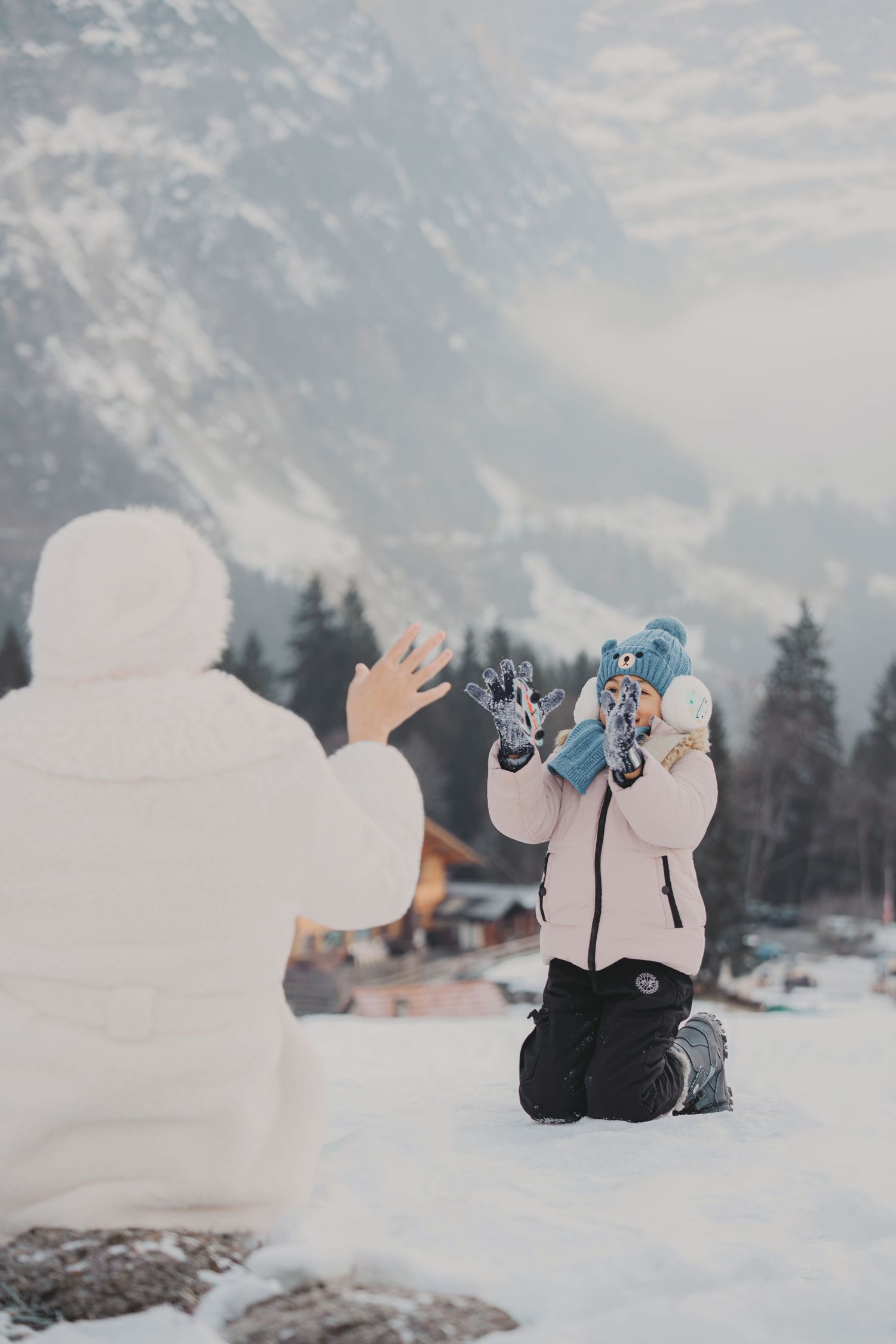 Young girl in blue hat kneeling in snow playing with mother in white coat against mountain backdrop.