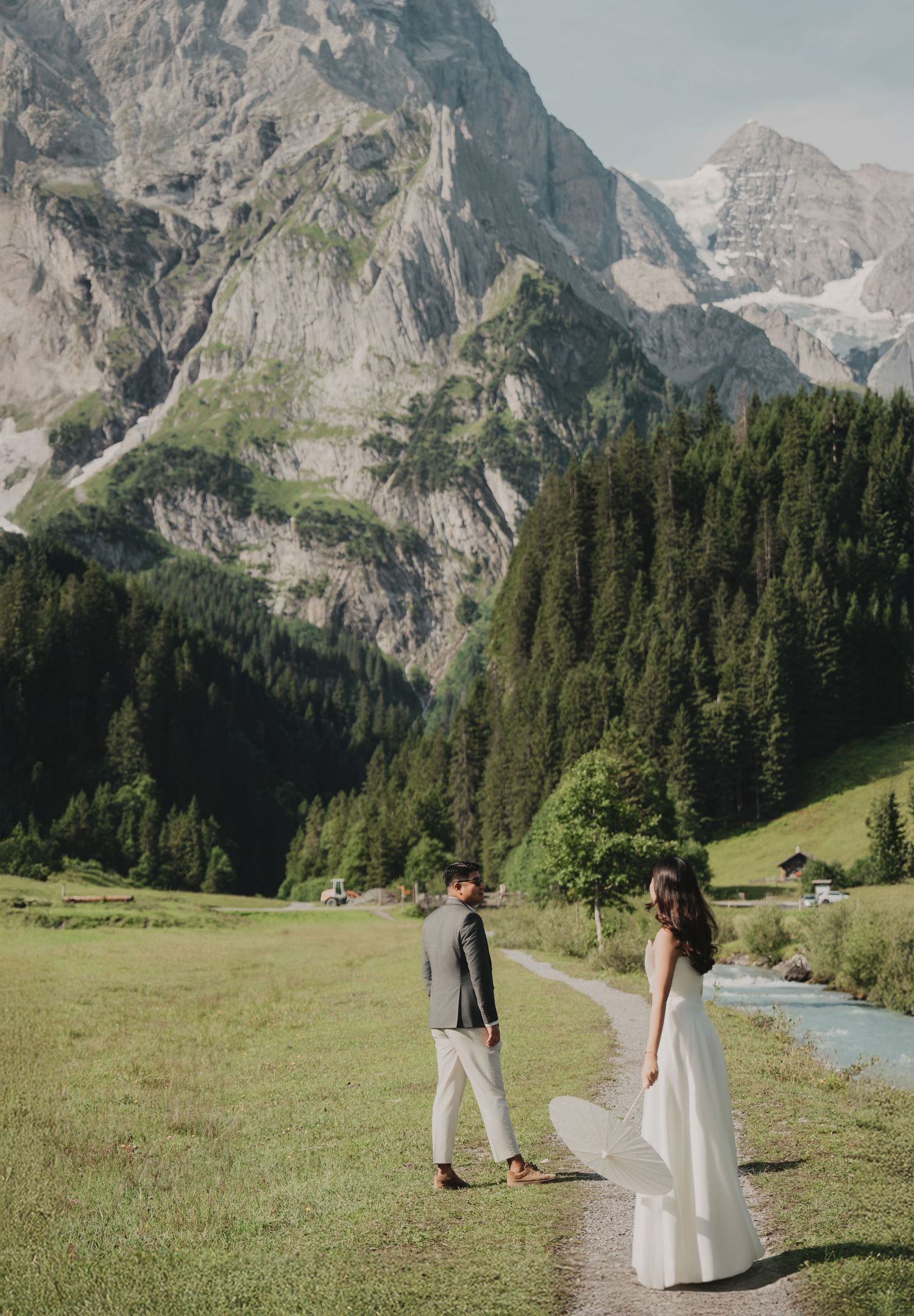 Bride and groom walking apart on a green meadow path with dramatic rocky mountain cliffs behind.