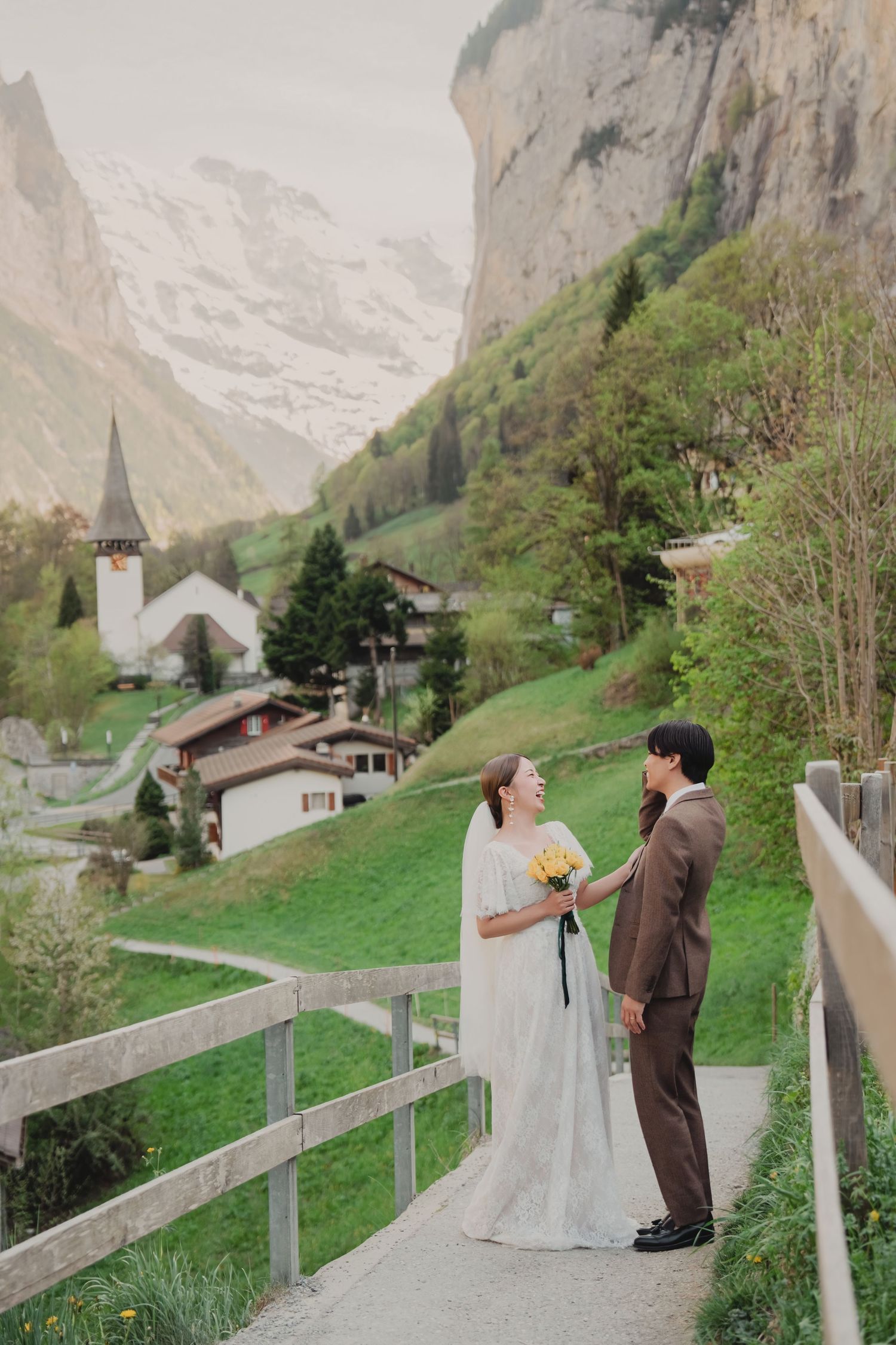 Wedding couple holding yellow bouquet on bridge overlooking scenic Lauterbrunnen valley church.