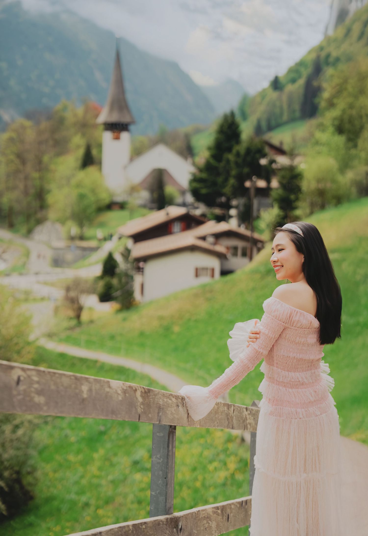 Smiling Asian woman in pink off-shoulder dress standing by fence with Swiss village church and green hills behind.
