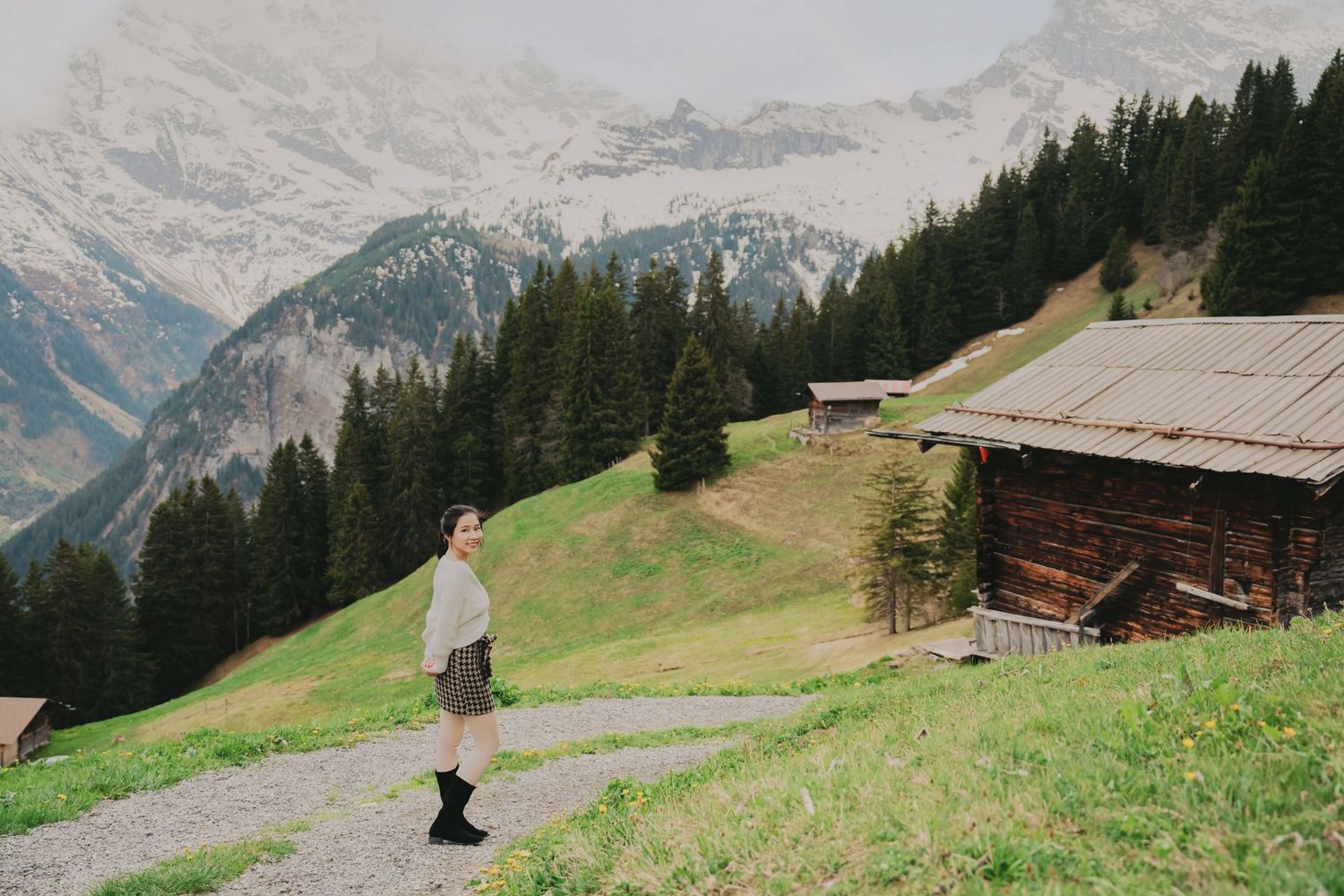 Woman in plaid skirt walks alpine path with snow-capped mountains and wooden chalet in Swiss Alps.