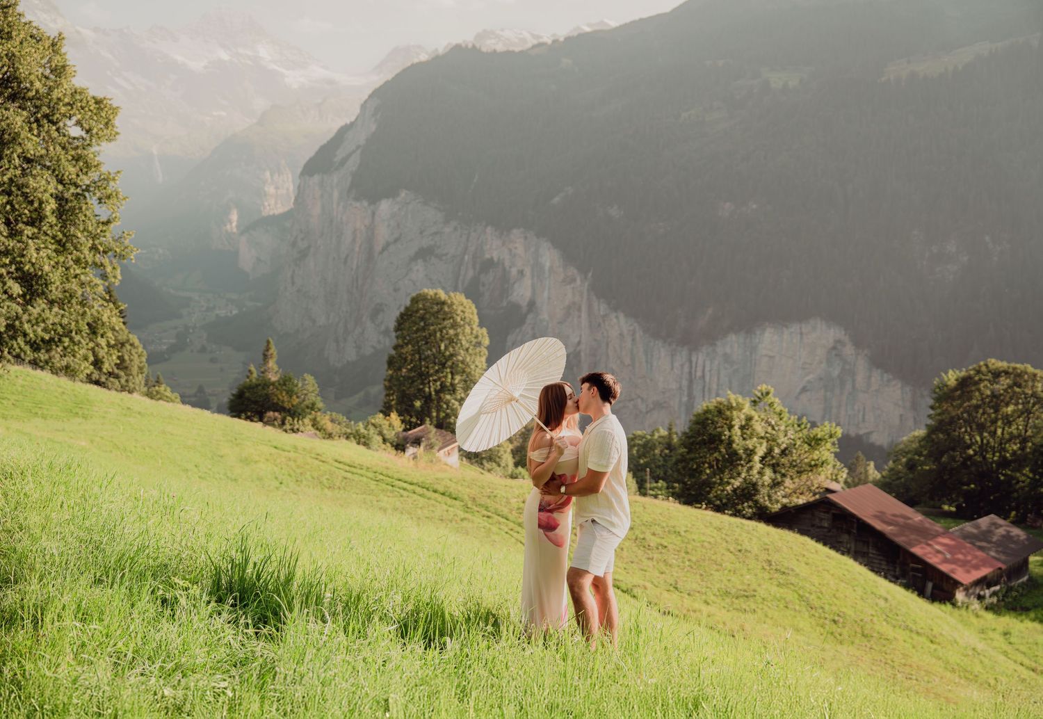 Romantic couple sharing kiss under white umbrella on lush green Swiss Alps hillside meadow.