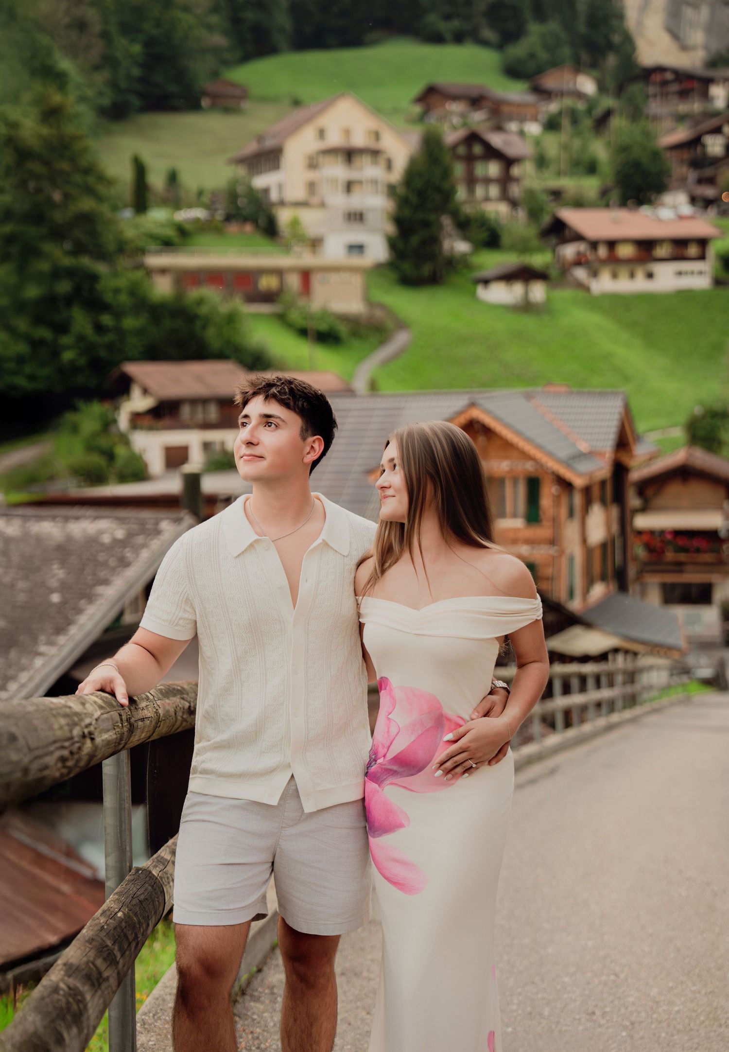 Couple standing on bridge admiring views of traditional Swiss alpine village on green hillside.