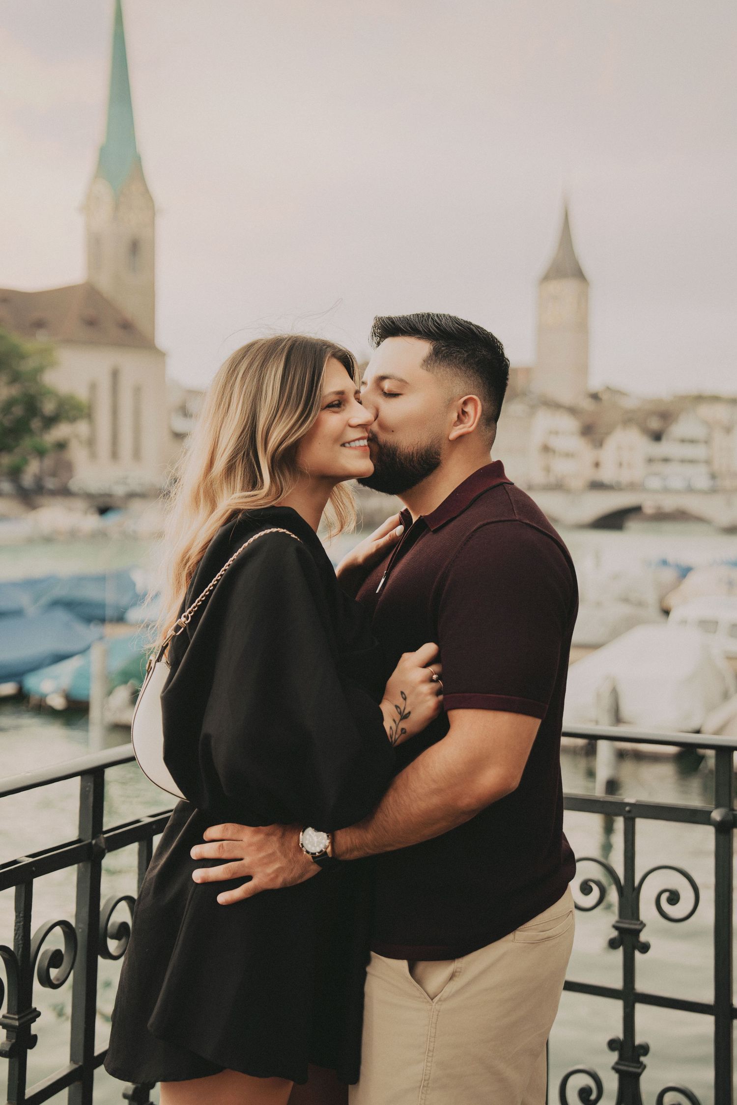 Man kissing womans cheek tenderly on a balcony with Zurich waterfront and church spires behind.