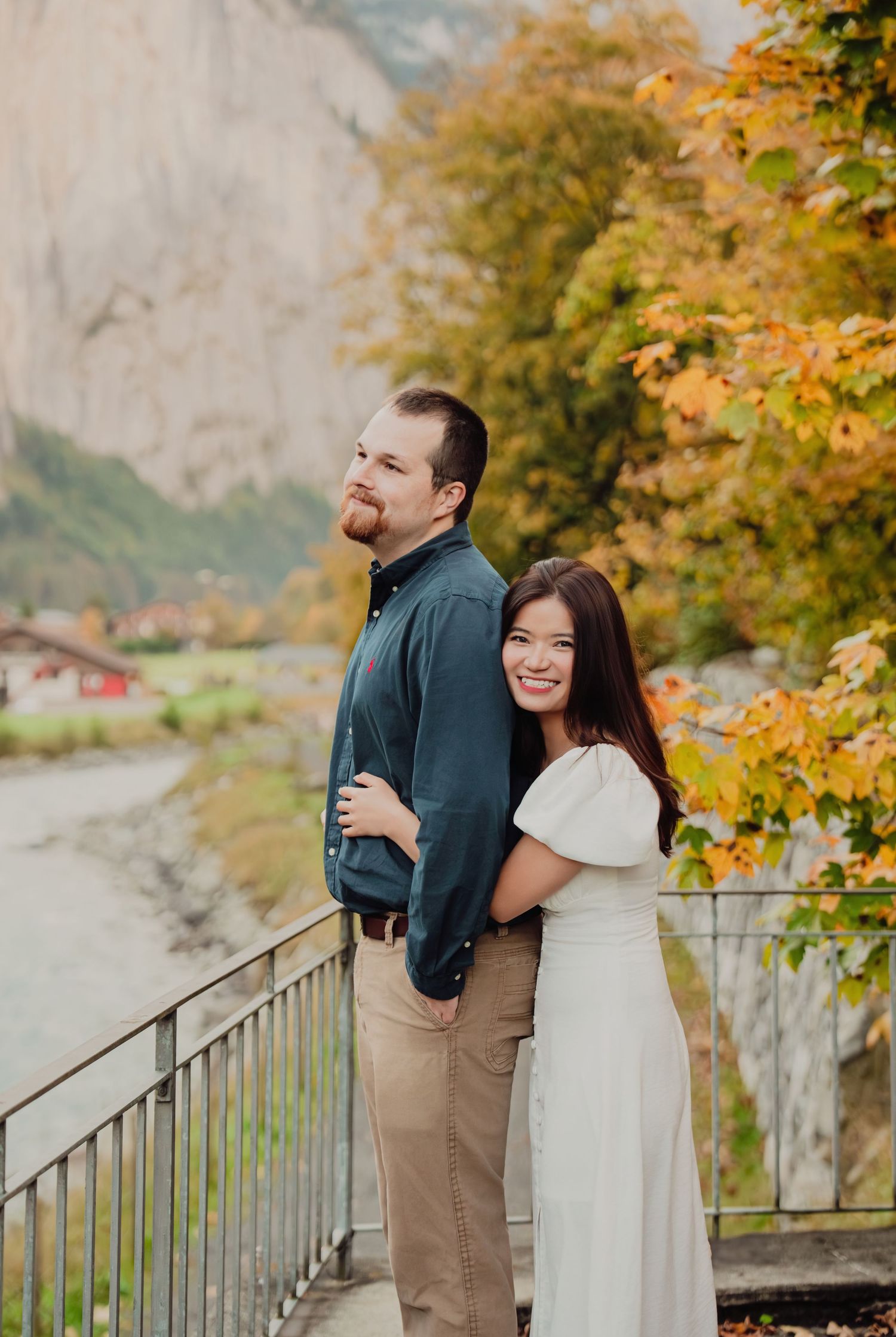 Couple posing back-to-back on a bridge with autumn foliage and mountain valley backdrop.