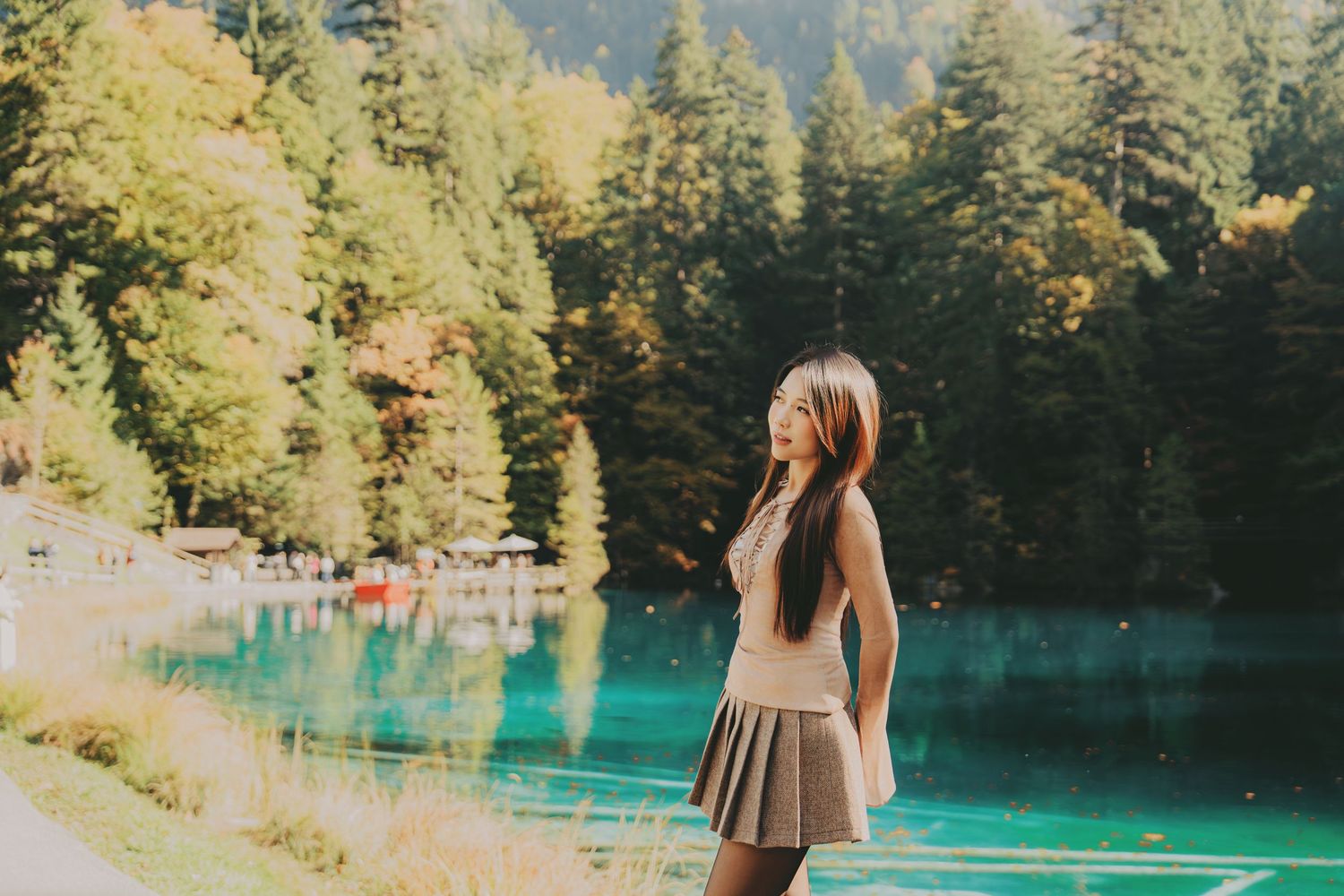 Young woman in beige top and gray skirt stands by a vivid turquoise lake in an autumn forest.