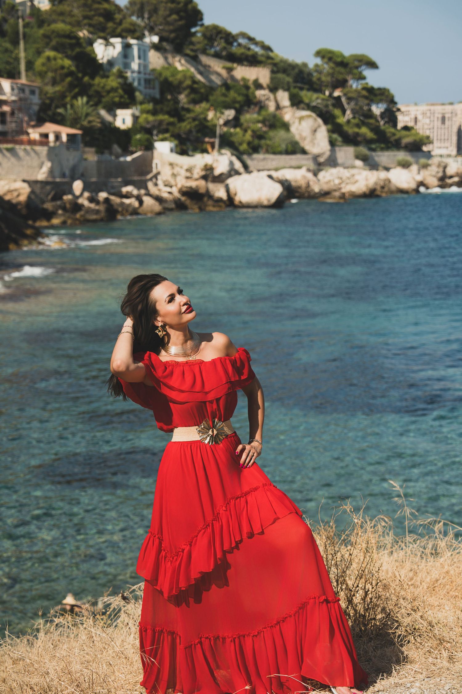 Elegant woman in flowing red off-shoulder dress posing on coastal cliff with blue Mediterranean sea.