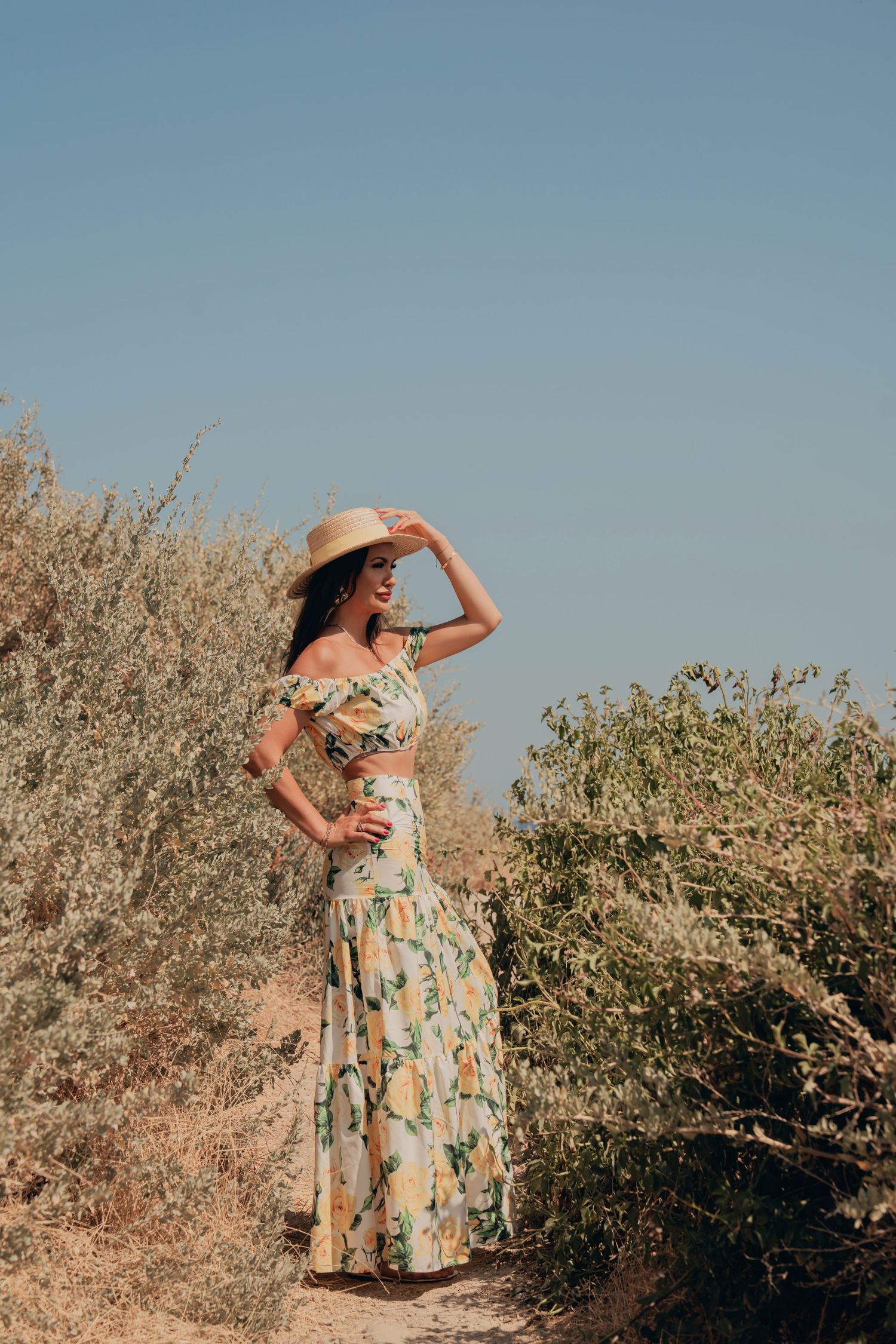 Woman in floral off-shoulder dress and straw hat standing among dry shrubs under blue sky.
