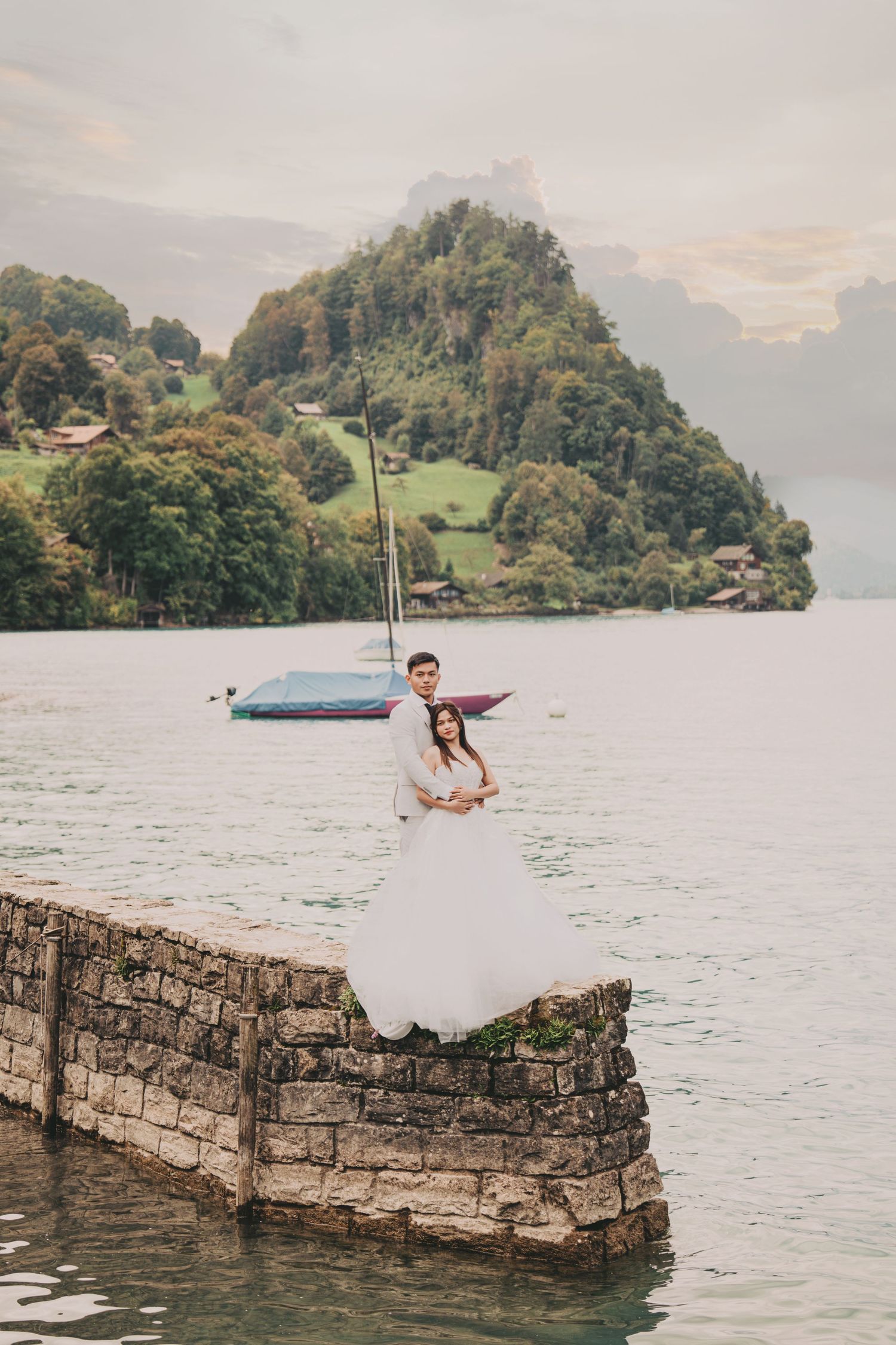 Groom embracing bride on stone dock with moored sailboat and green alpine mountain scenery.