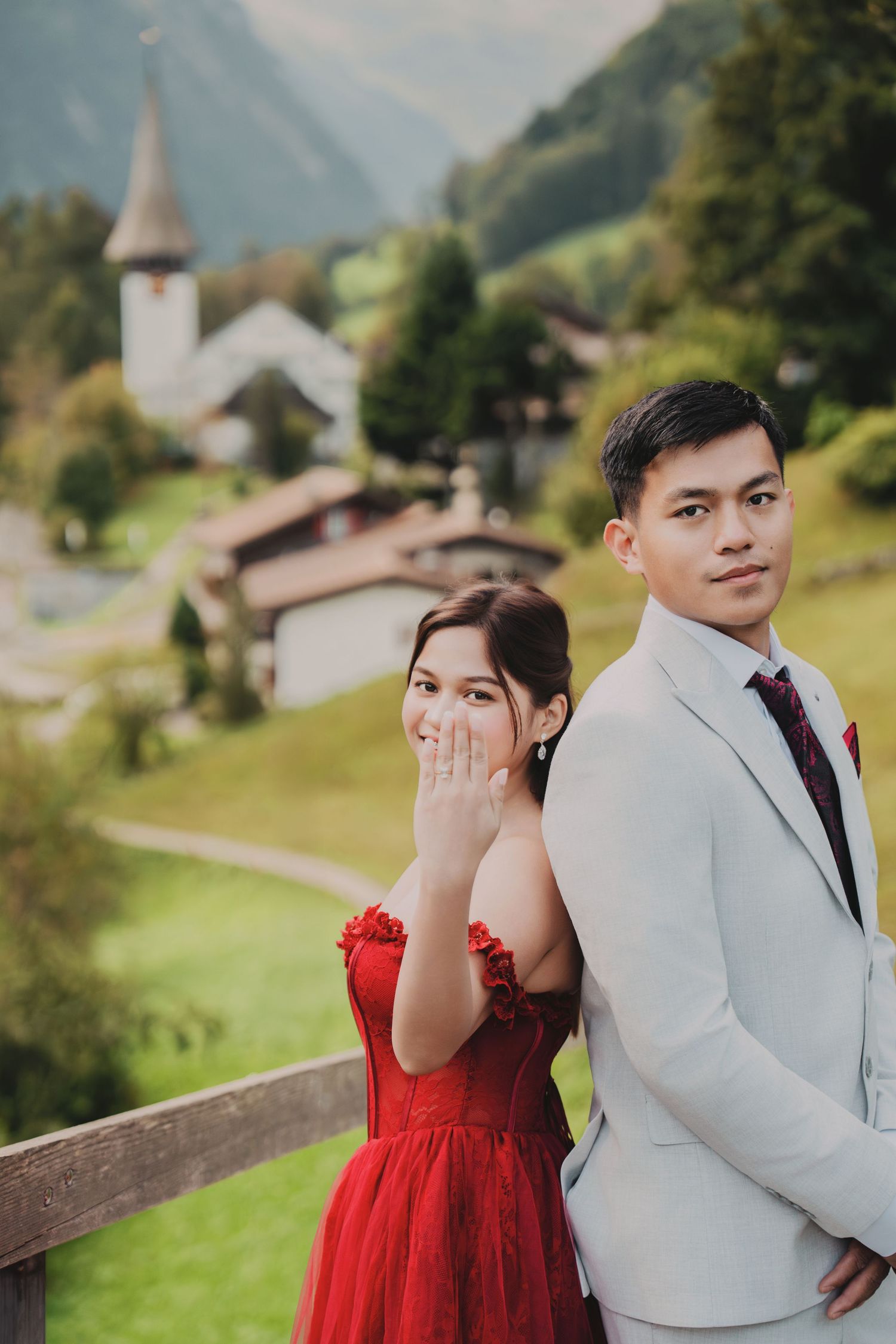 Engaged couple back-to-back showing diamond ring with charming Swiss village church in background.