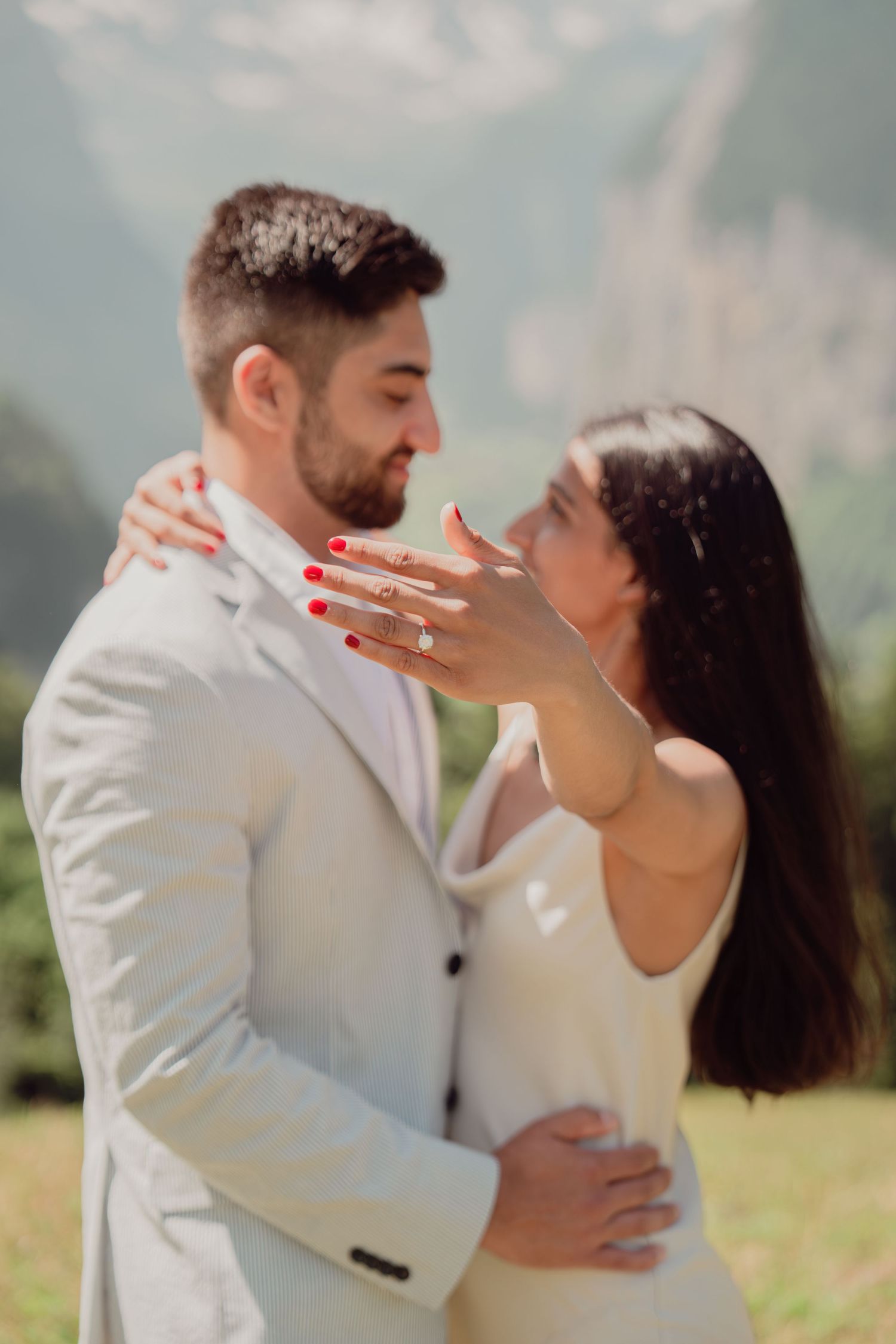 Romantic couple embracing closely as woman shows engagement ring with mountain scenery in the background.