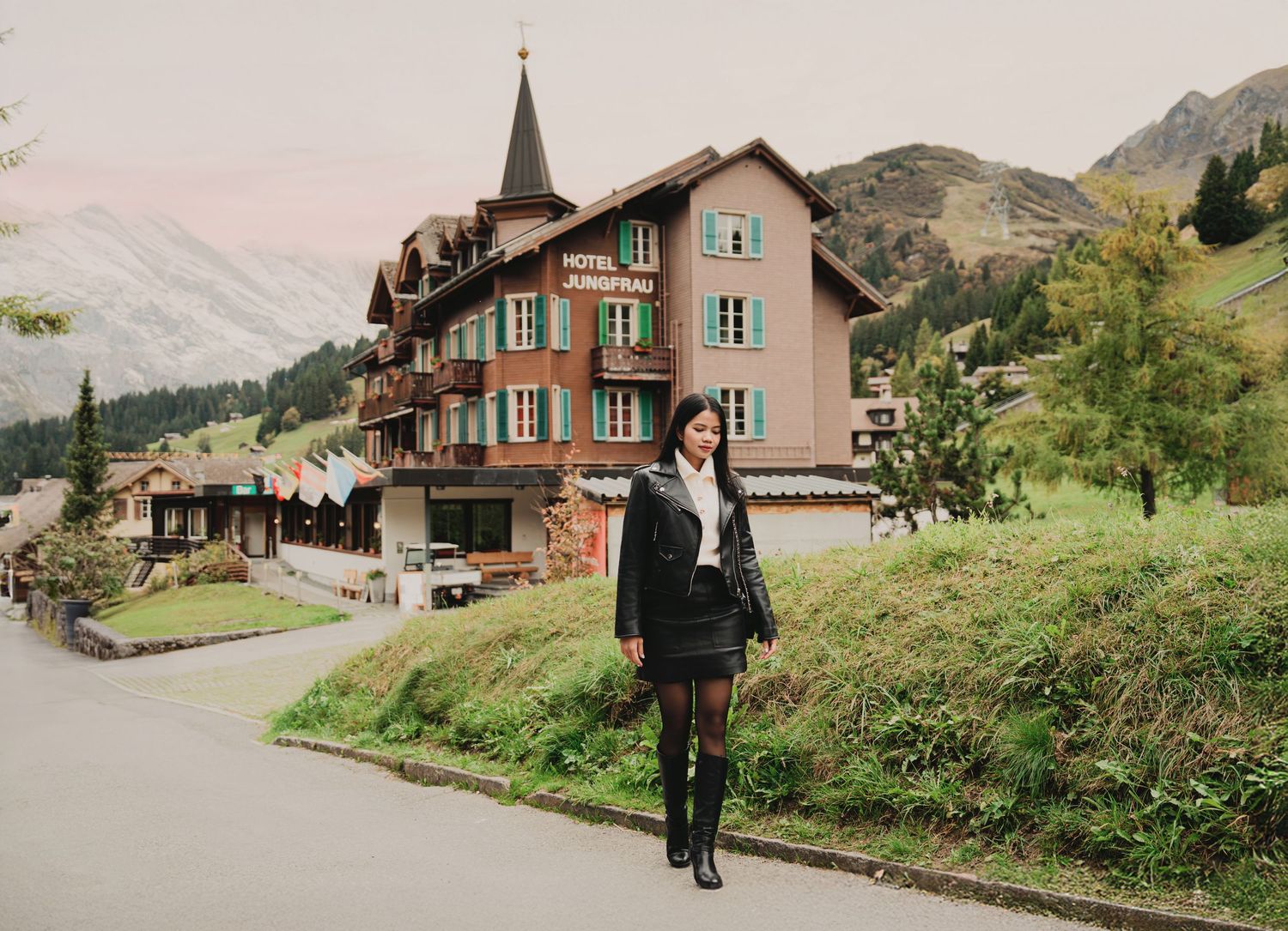 Young woman in black jacket and mini skirt stands before Hotel Jungfrau in Swiss Alpine village.