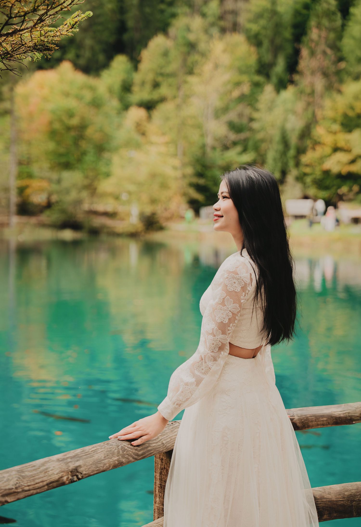 Smiling woman in elegant white lace dress poses by turquoise lake with autumn trees.