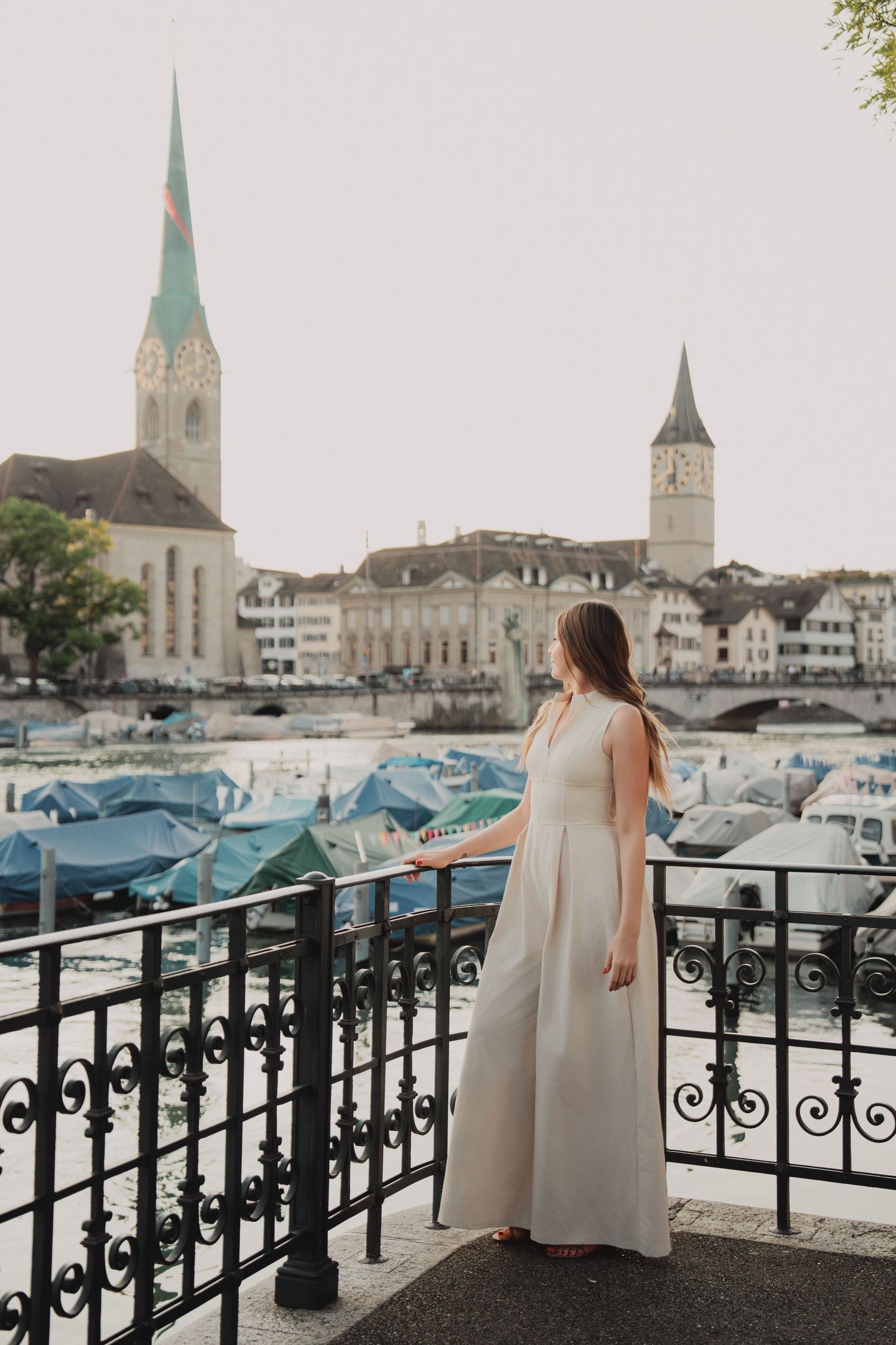 Woman in white gown standing at iron railing overlooking Zurich river and church spires.