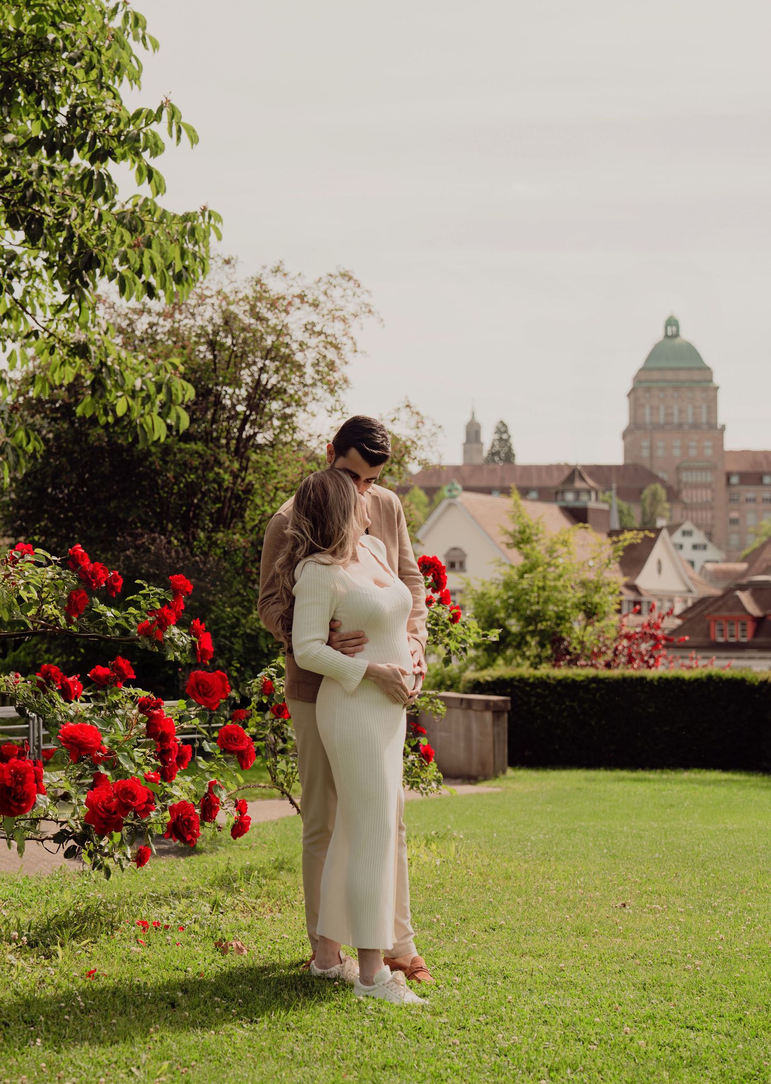 Couple sharing an intimate embrace near blooming red roses with a European city skyline backdrop.