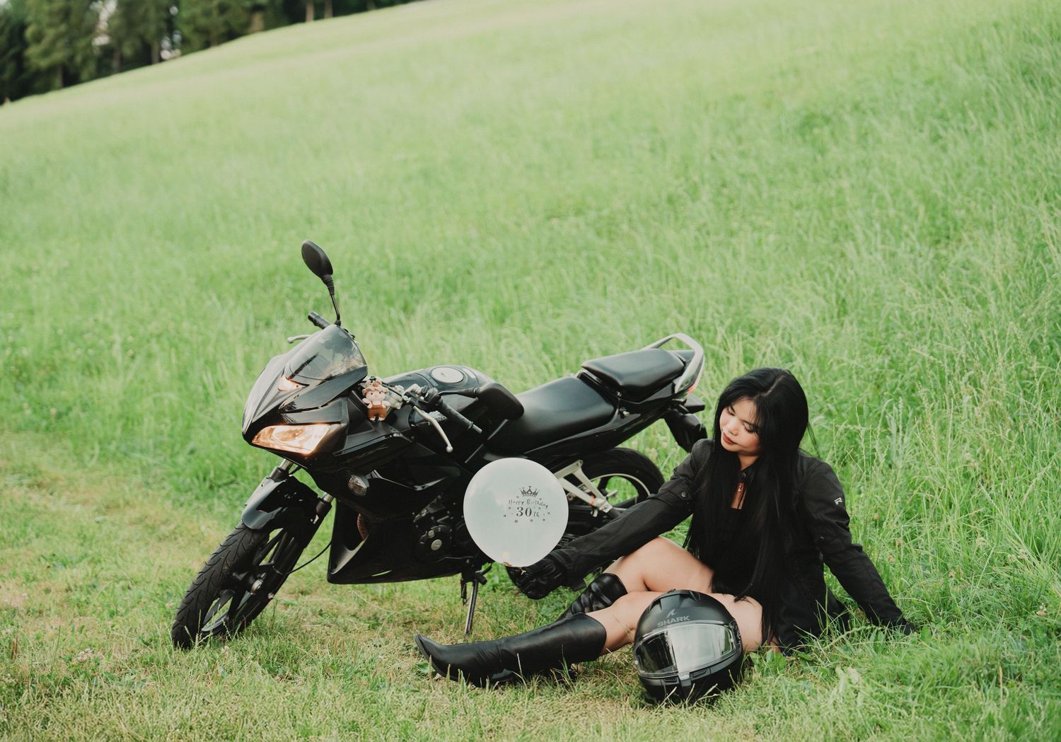 Young woman in black outfit posing with a black motorcycle on a lush green field.