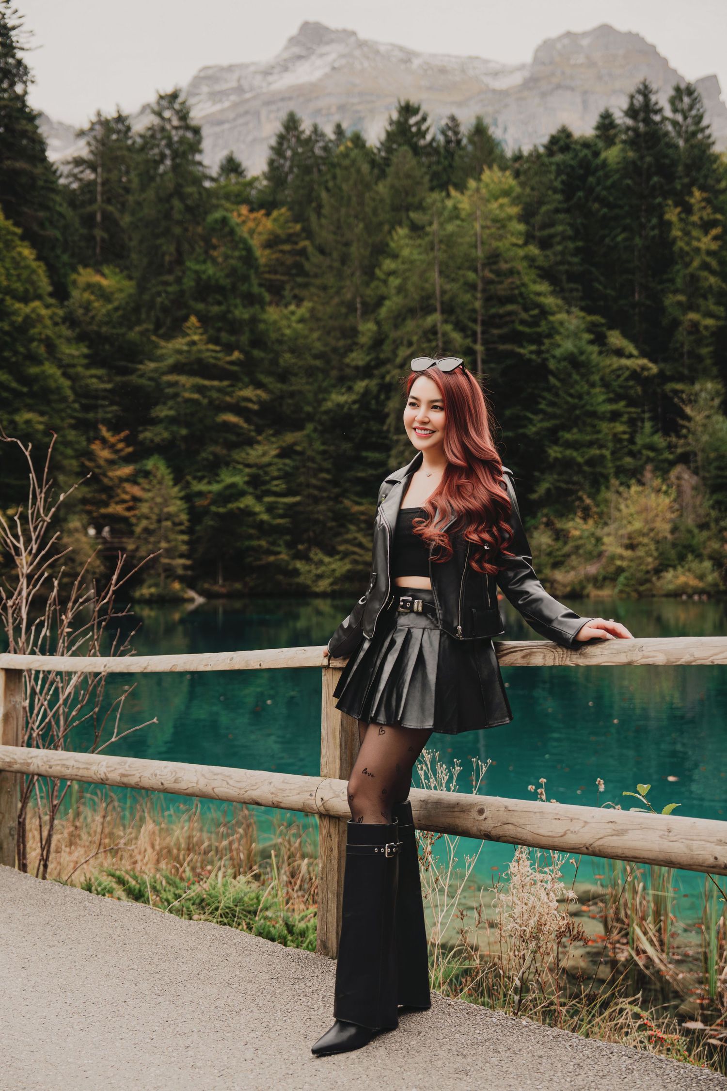Stylish woman in black leather jacket and mini skirt standing by turquoise lake with mountain views.