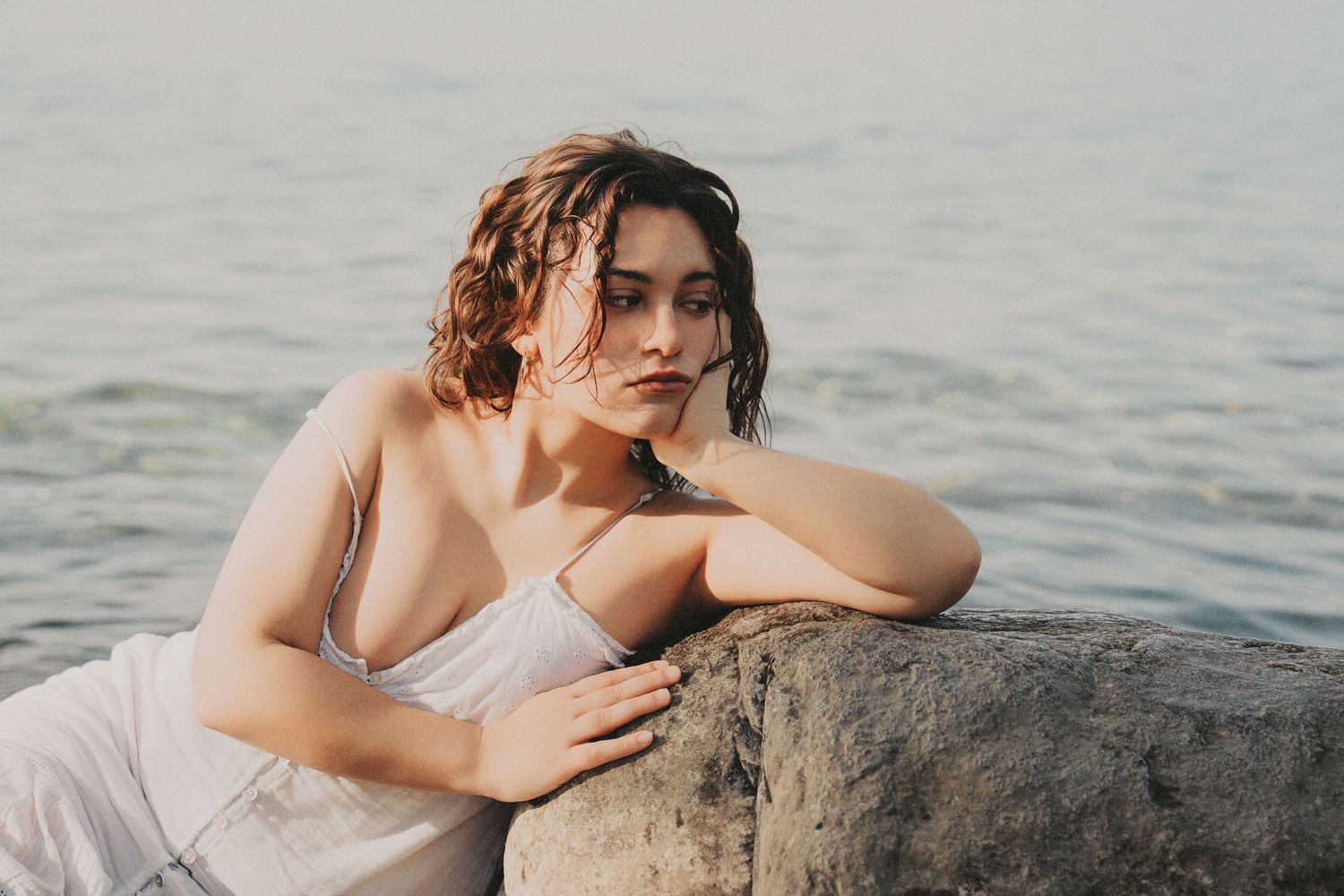 Young woman in white dress leaning on rock by the sea, looking pensive.