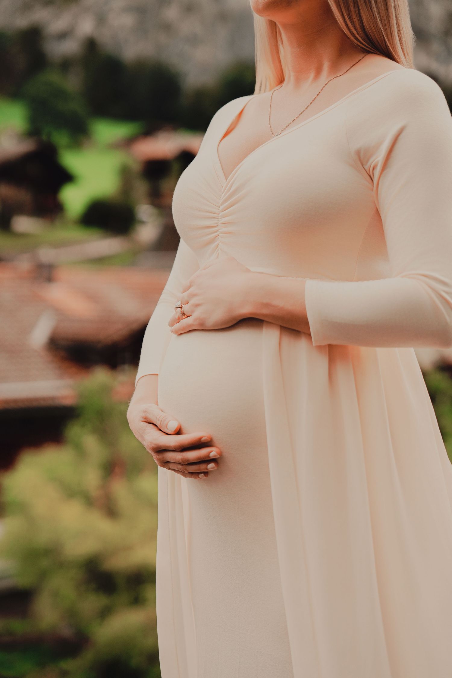 Close-up of a pregnant woman in a blush dress cradling her baby bump with a Swiss village backdrop.