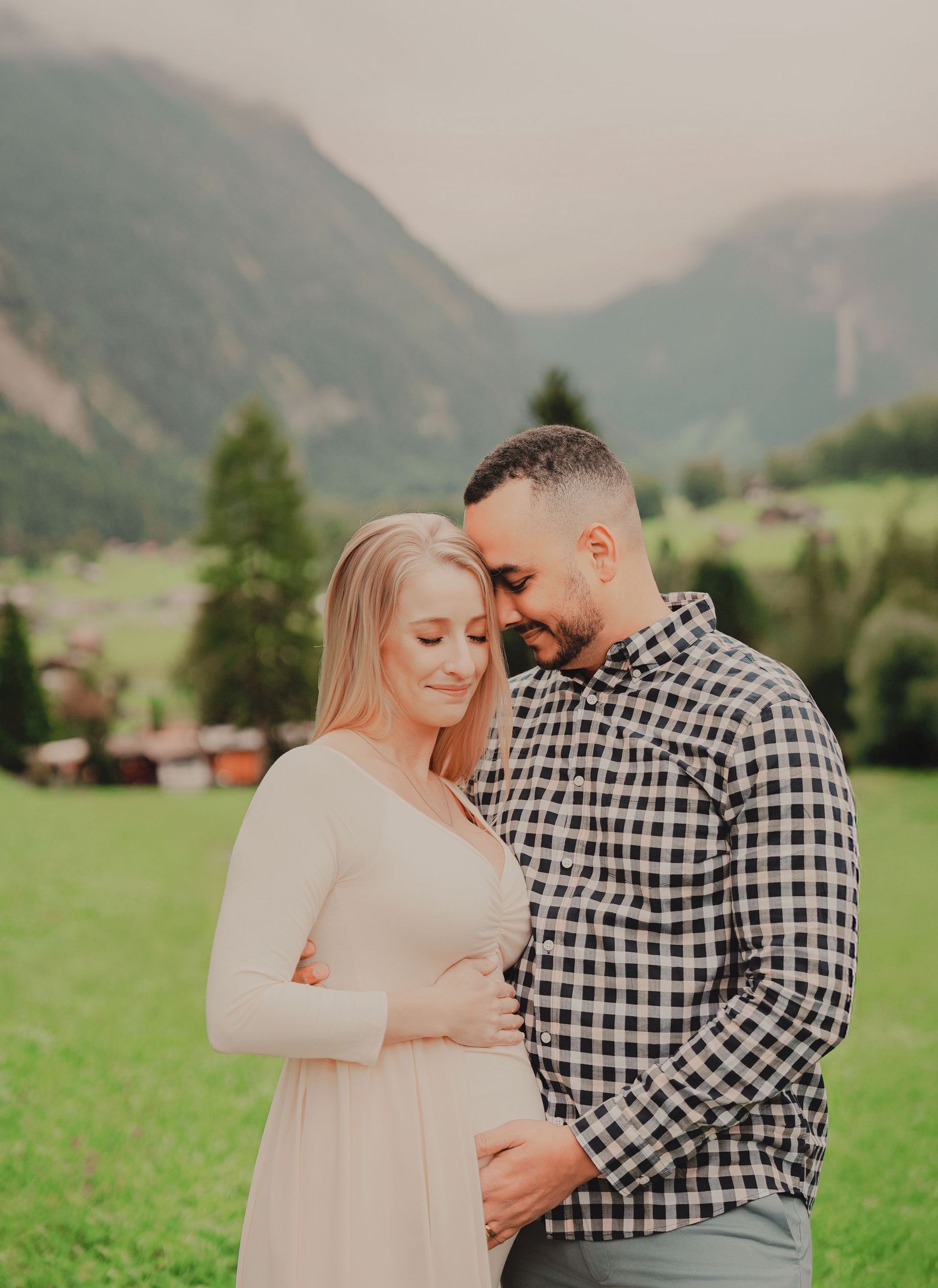 Tender expecting couple embracing in soft light with lush green Swiss alpine meadow and mountains behind.