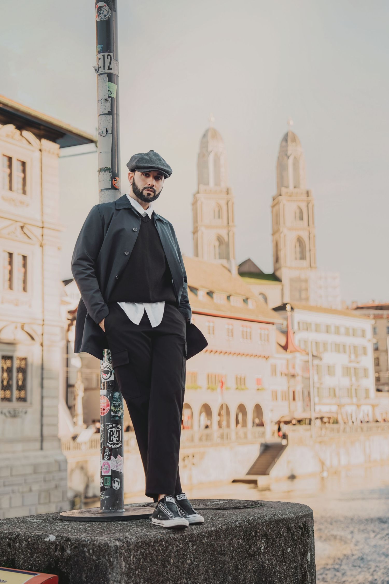 Bearded man in dark coat and flat cap standing on a stone platform near Grossmunster church in Zurich.