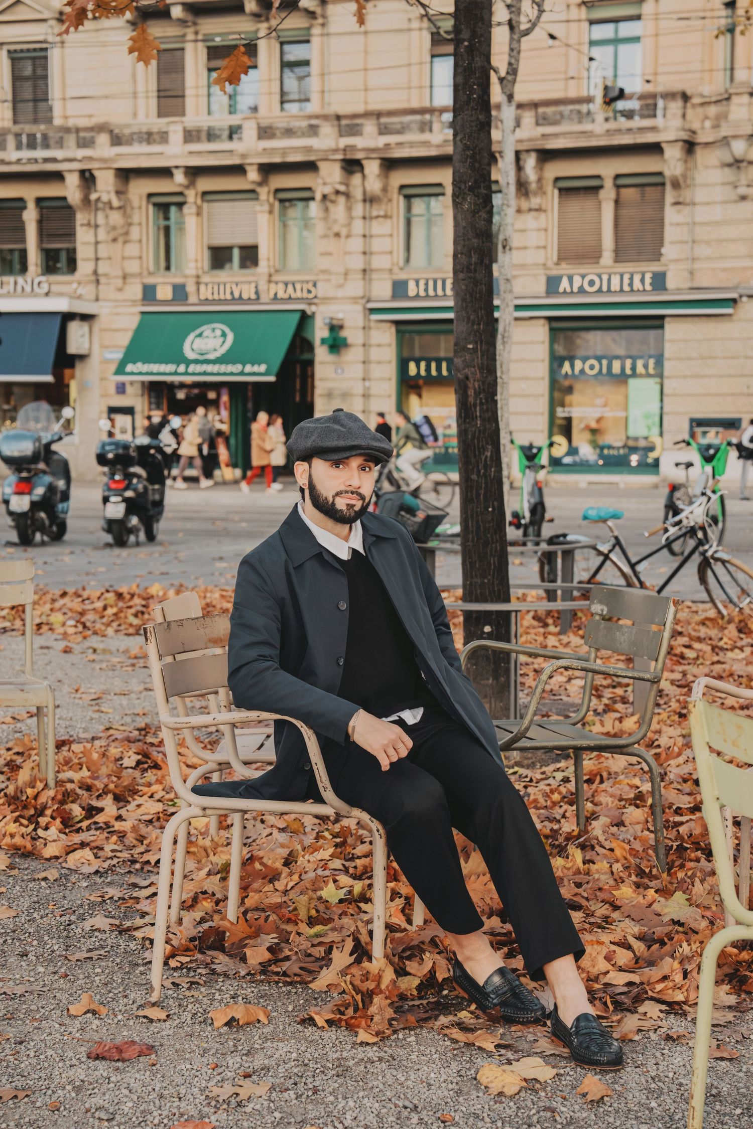 Stylish man in black coat and flat cap sitting on chair among autumn leaves in city.