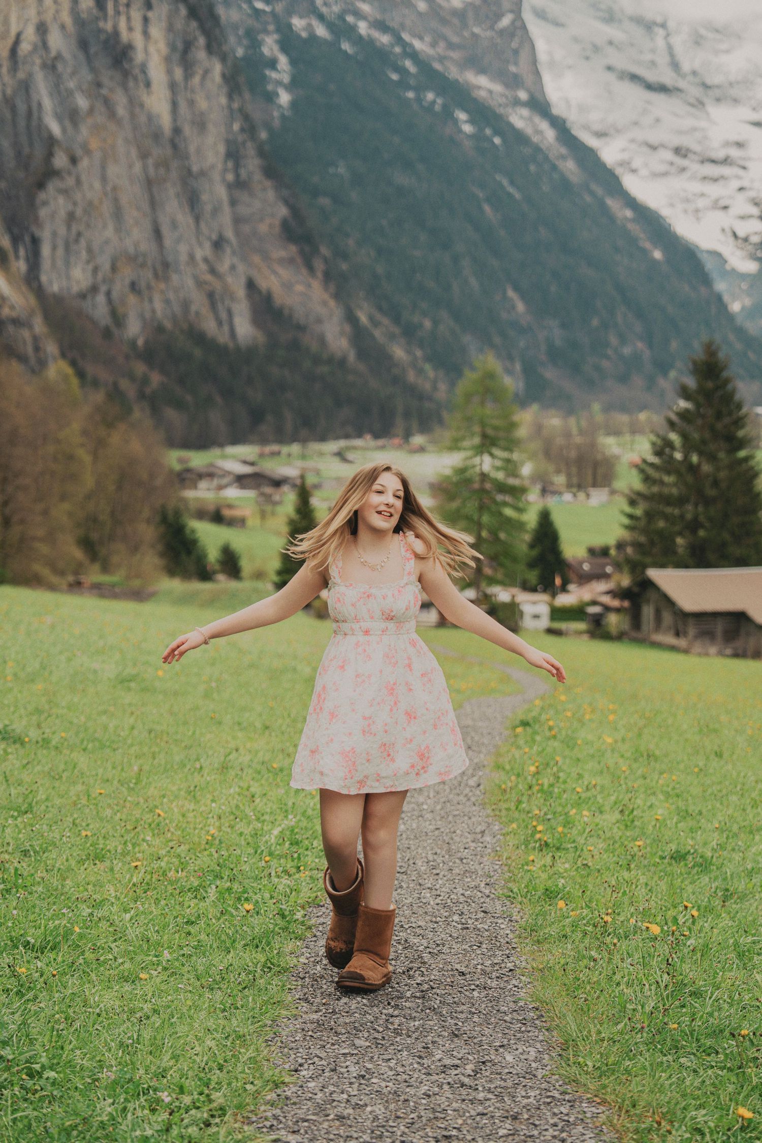 Carefree young woman in floral dress spinning with arms wide open on a scenic gravel path in Swiss Alps.