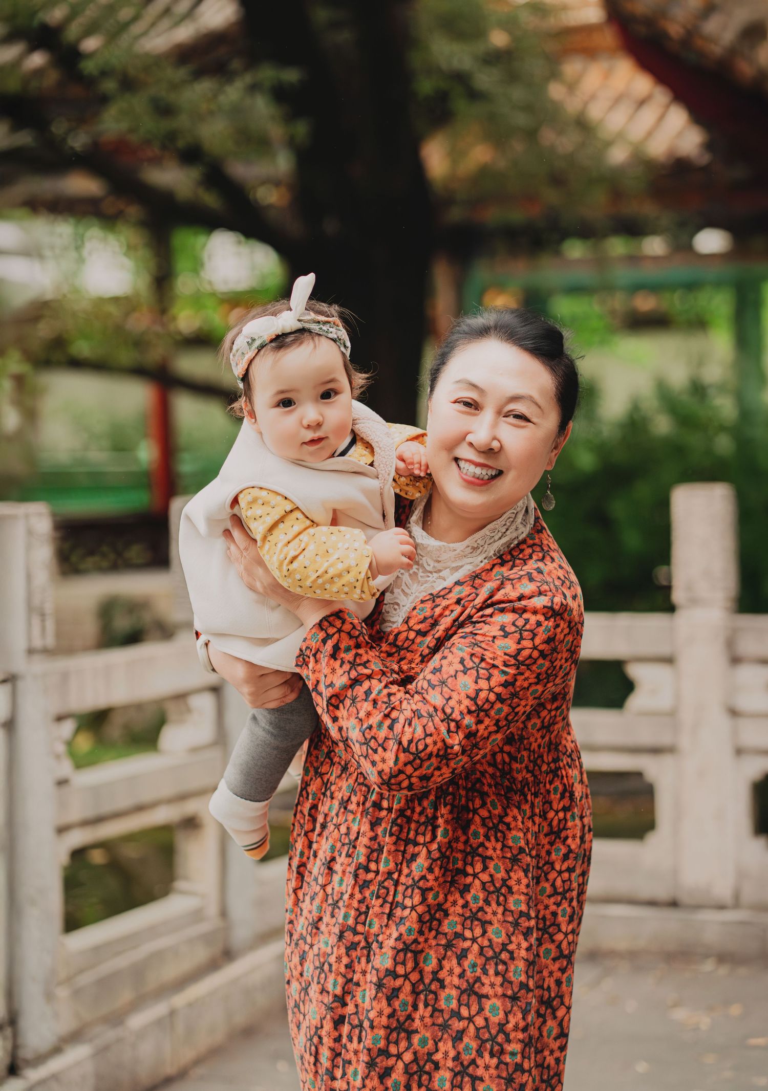 Grandmother in floral dress joyfully holds baby girl in a traditional Chinese garden.