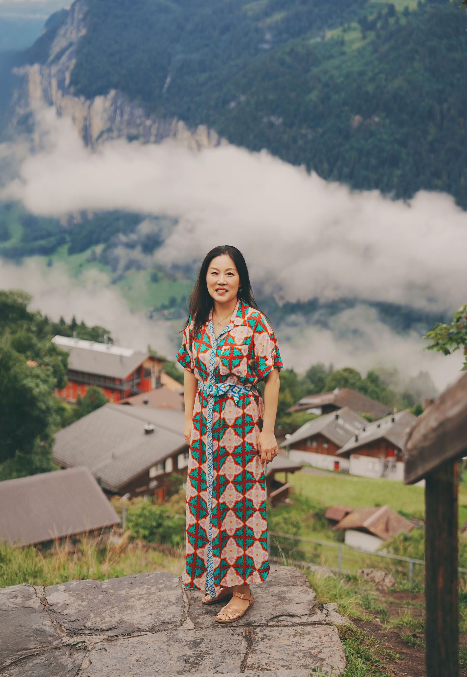 Woman in colorful patterned maxi dress standing on stone wall with Swiss Alpine village and misty mountains behind her.