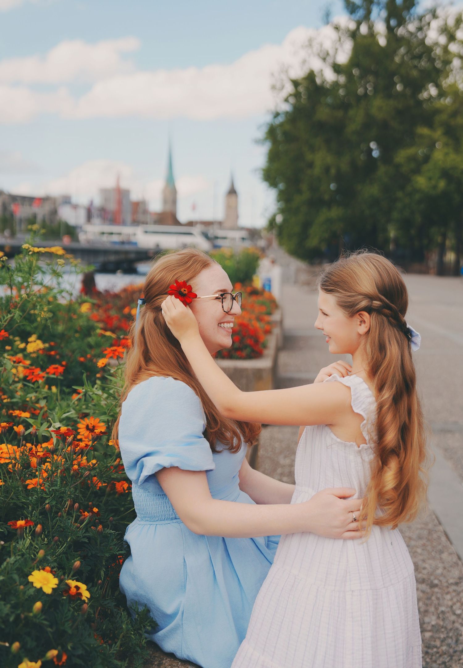 Woman placing red flower in girls hair by vibrant orange blooms near riverside cityscape.