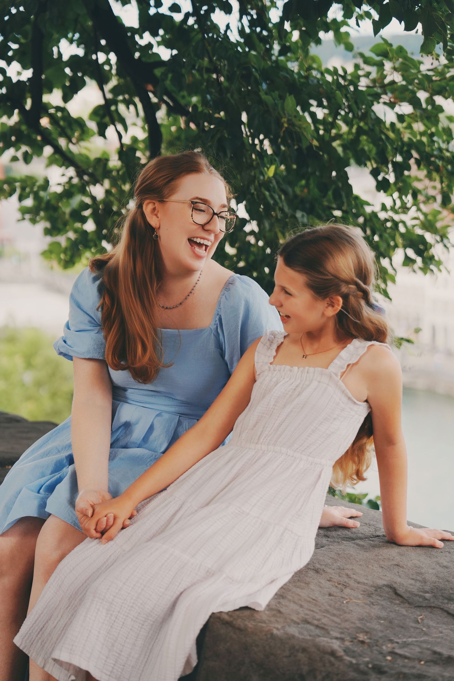 A laughing woman in a blue dress and a girl in white sit together on a stone ledge outdoors.