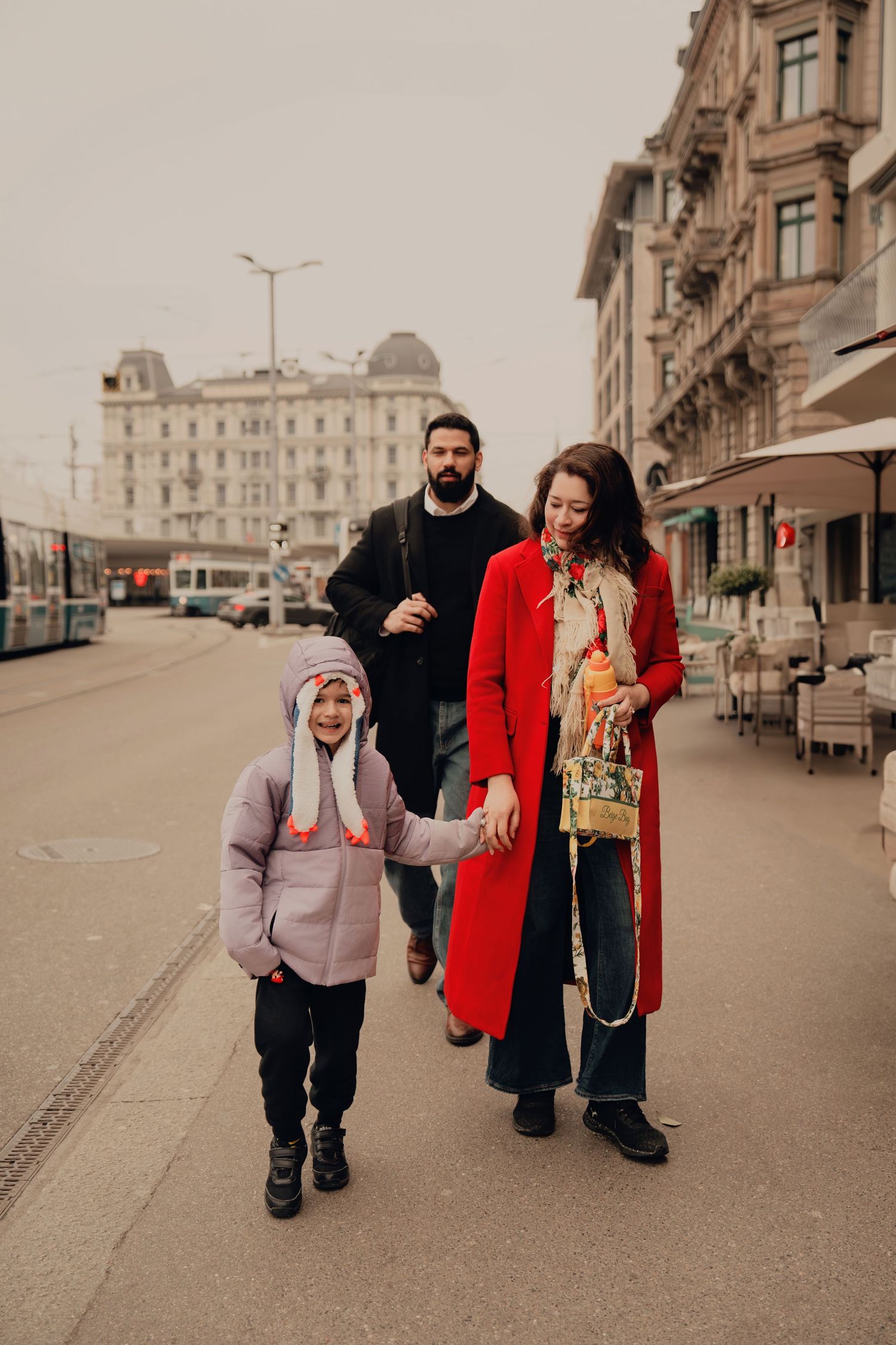Happy family strolling urban European street, child in purple jacket holding mother's hand in red coat.