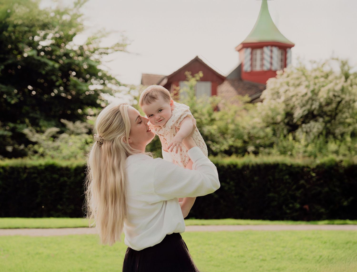 Blonde mother lifts laughing baby in air in lush green park with red building behind.