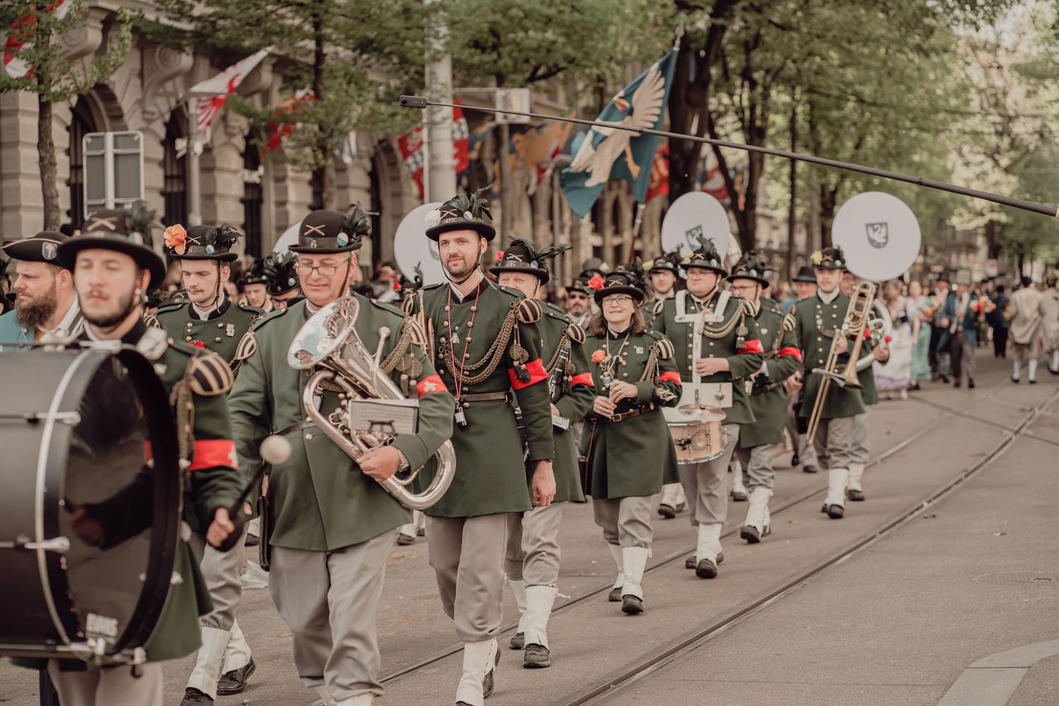 Marching band in green uniforms and traditional hats performing in a colorful street parade with flags overhead.