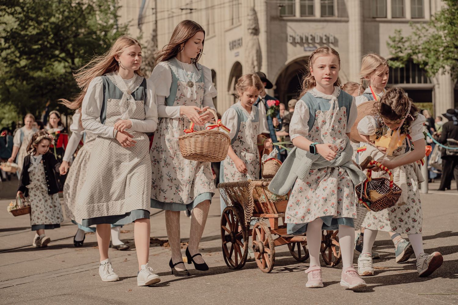 Girls in traditional folk costumes carrying wicker baskets march in a sunny outdoor parade.
