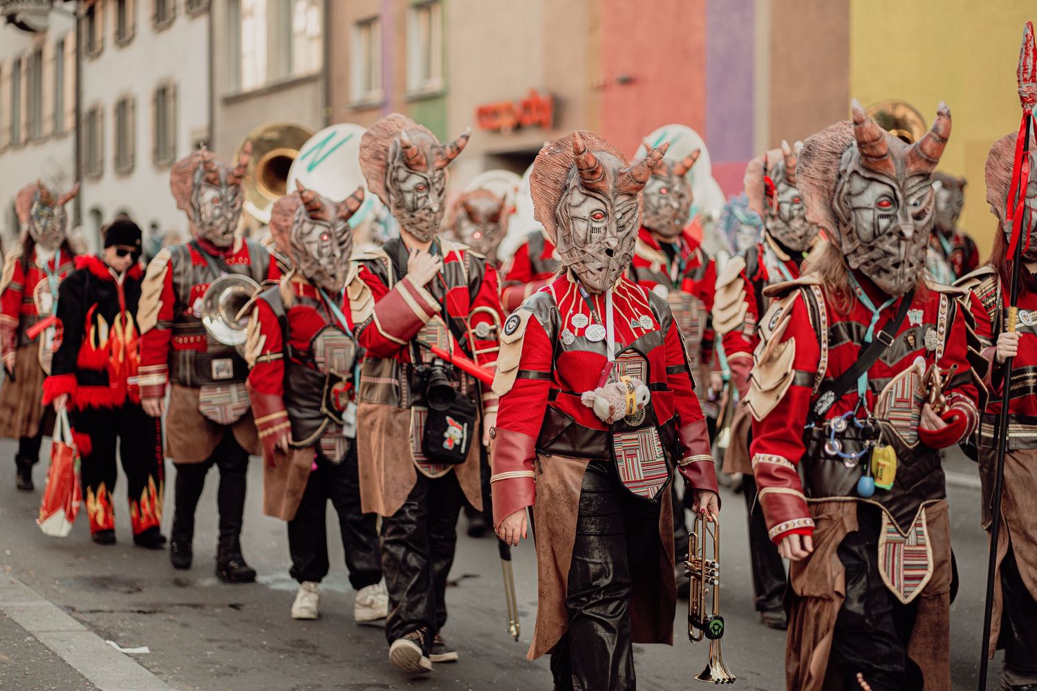 Performers in red jackets and horned demon masks marching through a street during a carnival parade.