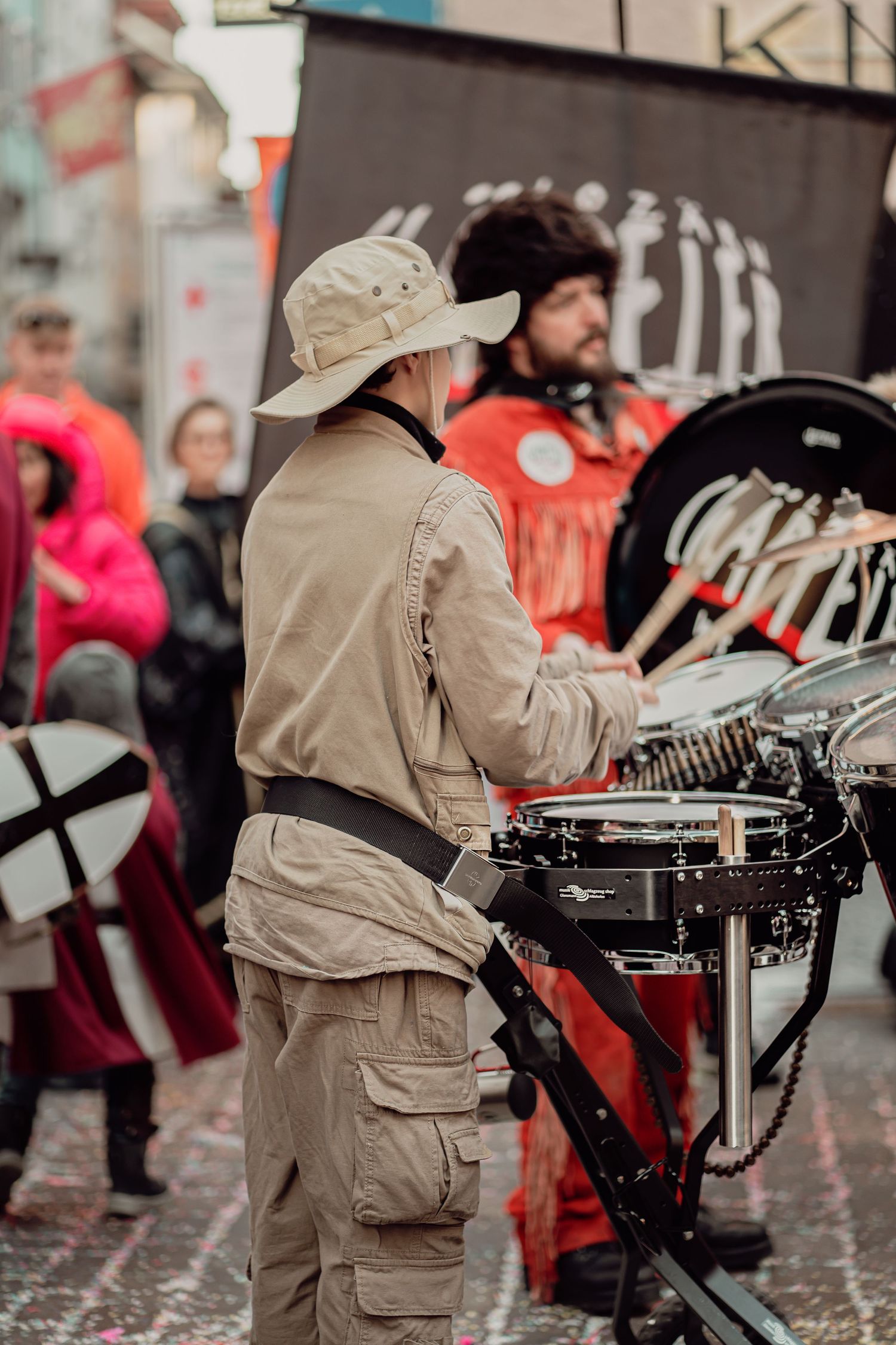 Drummer in beige uniform playing snare drums at an outdoor street performance or carnival event.