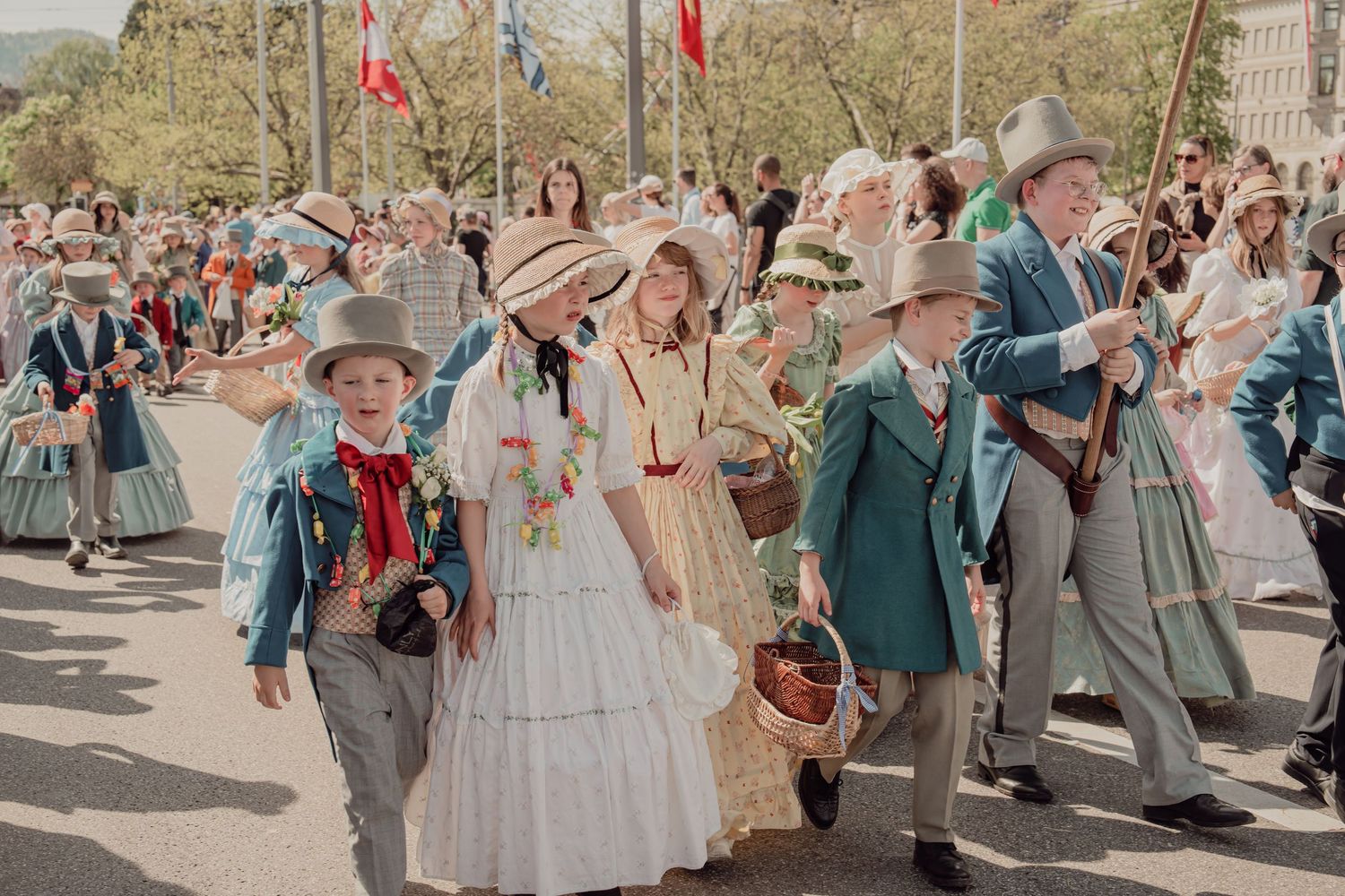 Crowd in Victorian-era costumes with straw hats marches in colorful spring parade under sunny skies.