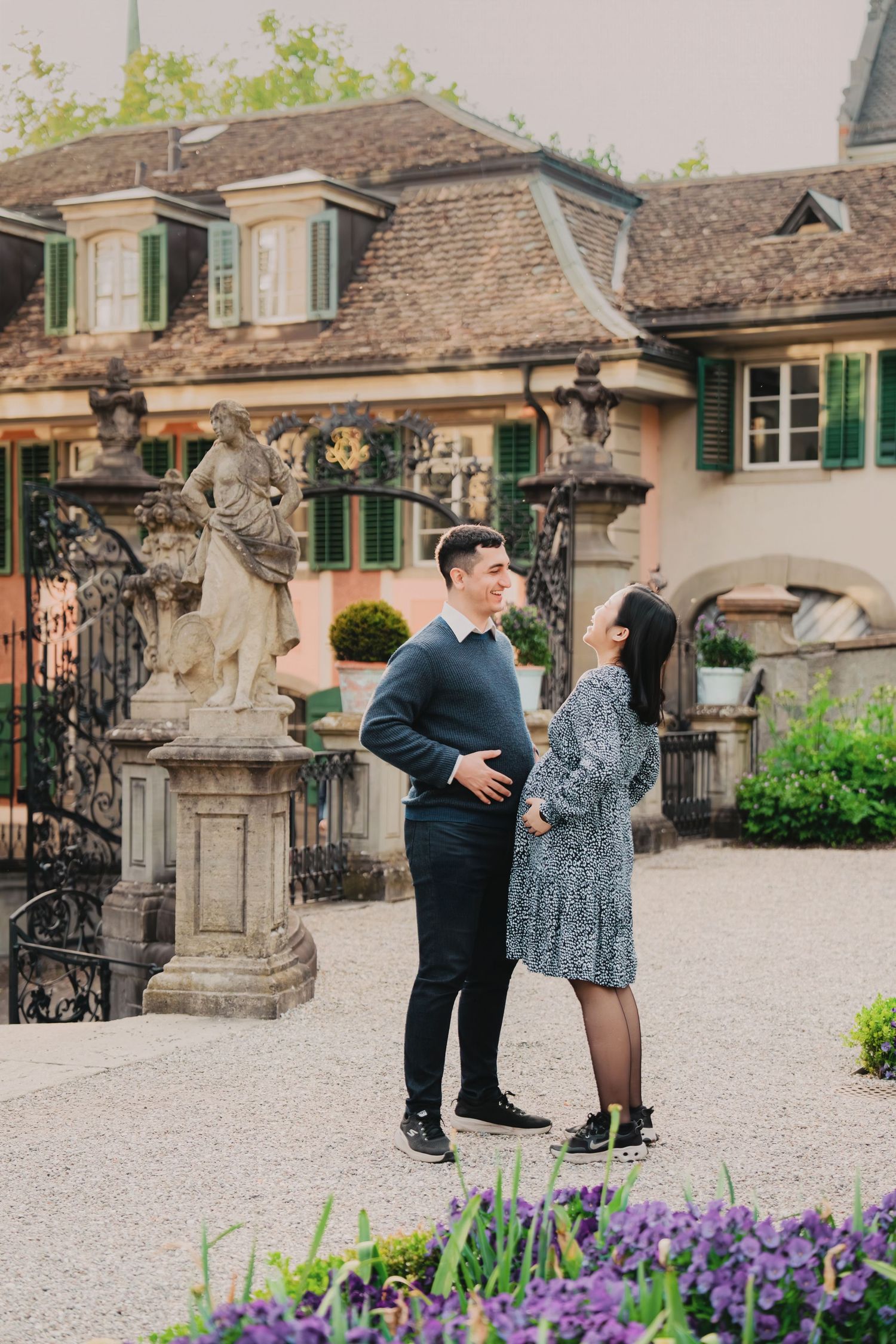 A couple shares a tender moment in an elegant European garden courtyard adorned with classical stone sculptures.