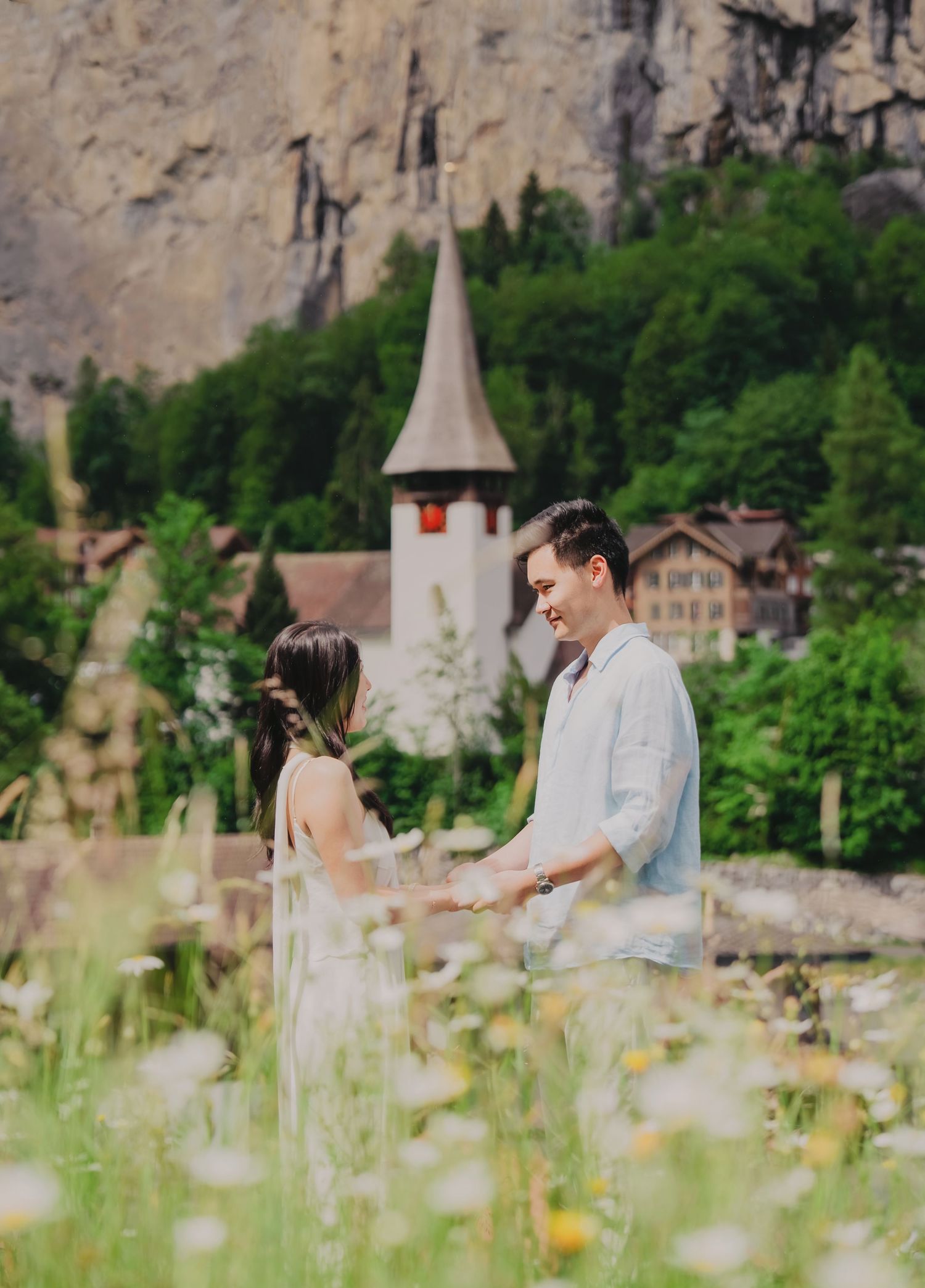 Couple holding hands in a wildflower meadow with a charming Swiss village church and rocky cliffs in the background.