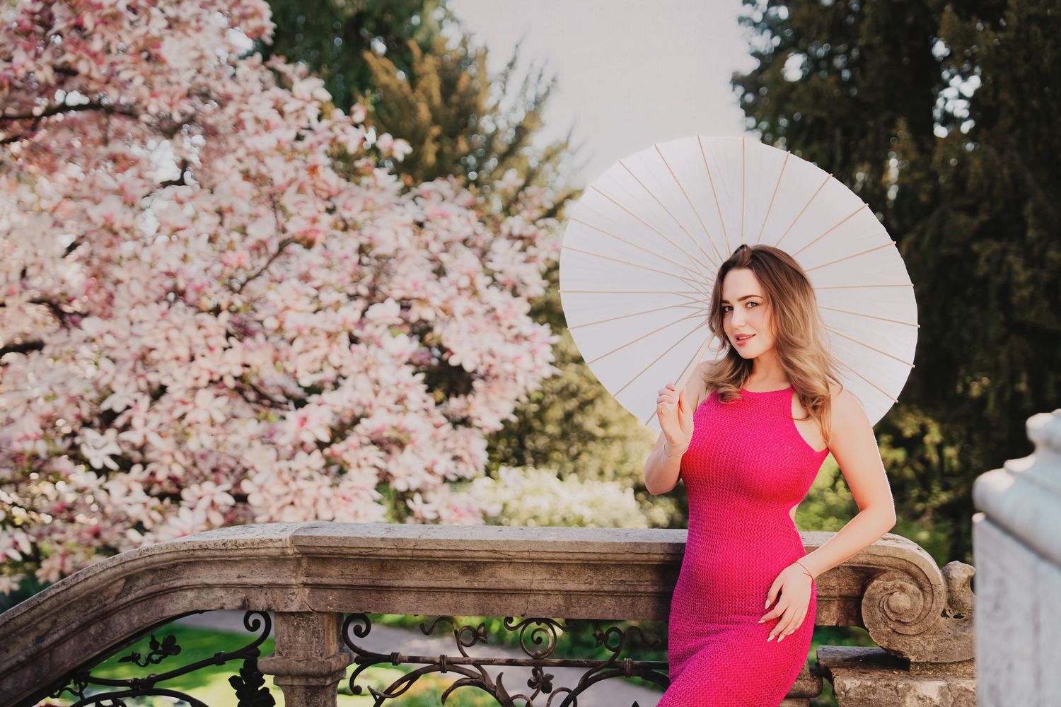 Woman in pink dress holding white paper umbrella near blooming magnolia trees on stone balcony.