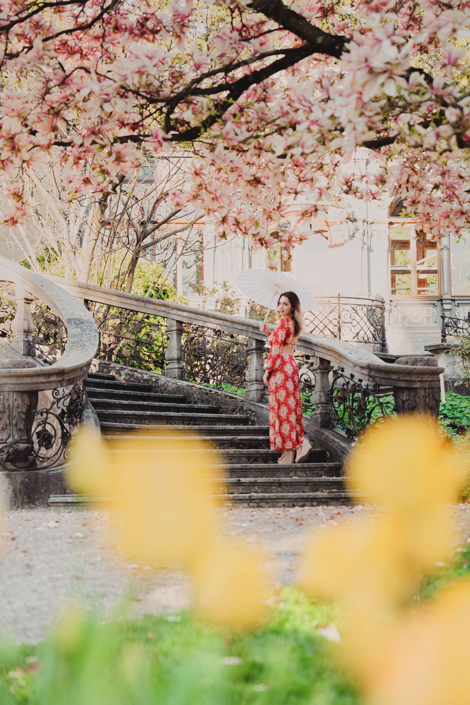 Woman in red floral dress standing on stone stairs under pink cherry blossom trees with yellow tulips.