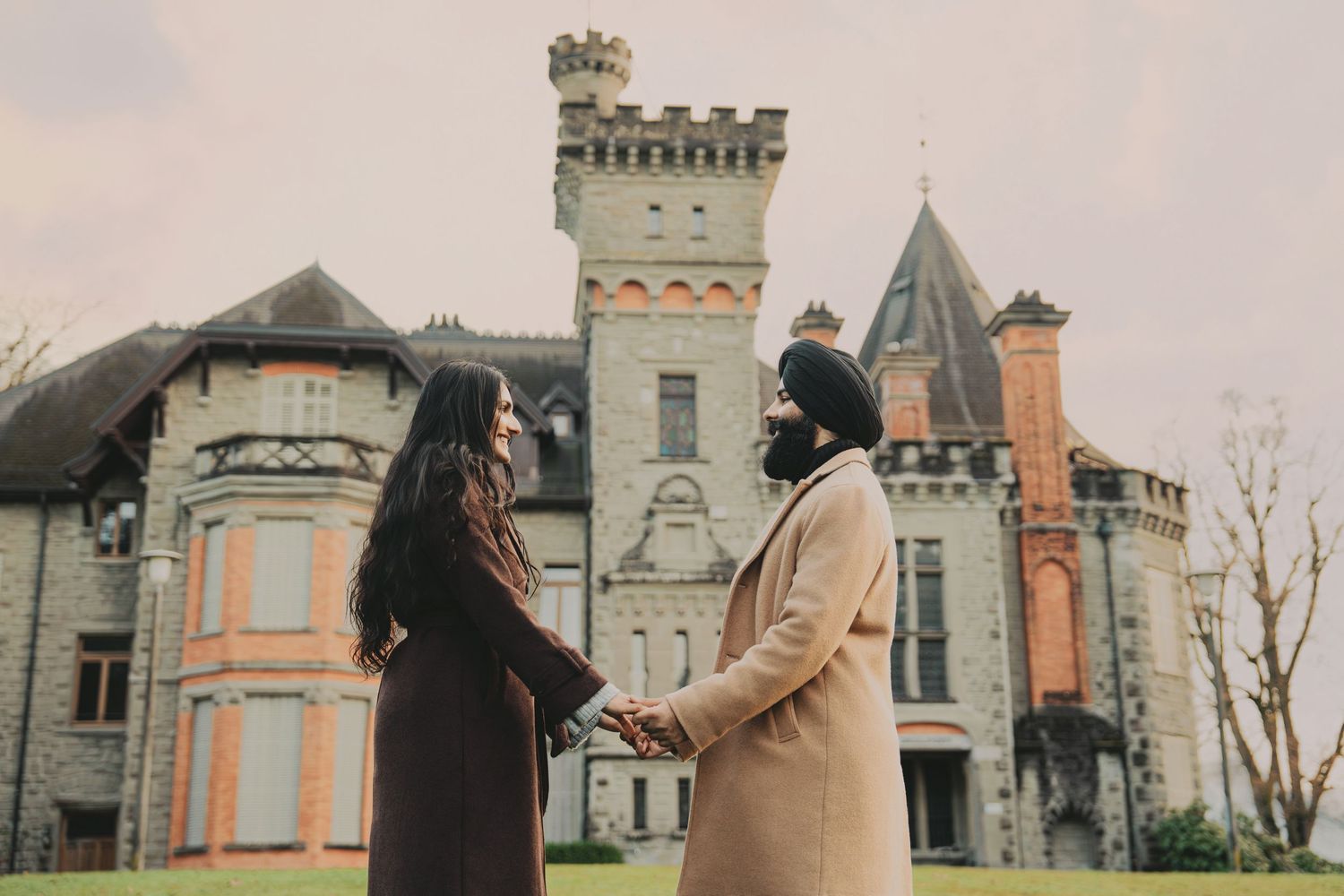 Couple holding hands facing each other in front of a grand castle during an autumn engagement photoshoot.