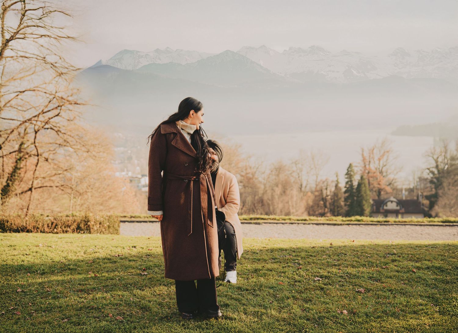 Sikh man kneeling before woman in brown coat during marriage proposal in sunny park with mountains.