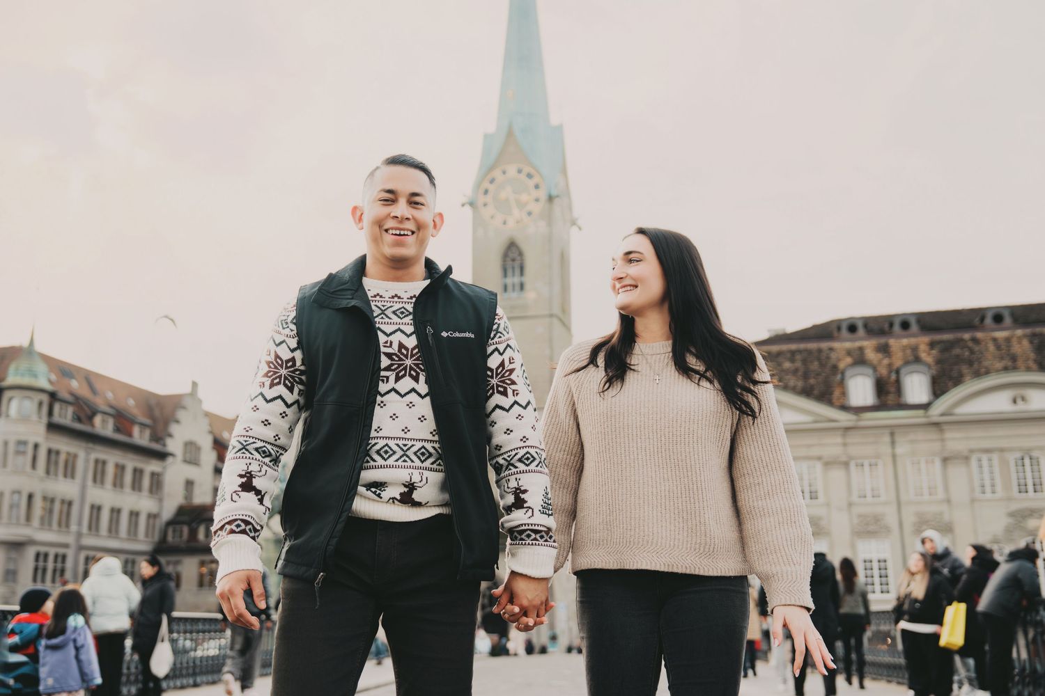 Joyful couple holding hands and laughing while walking near a Gothic church tower in Zurich.